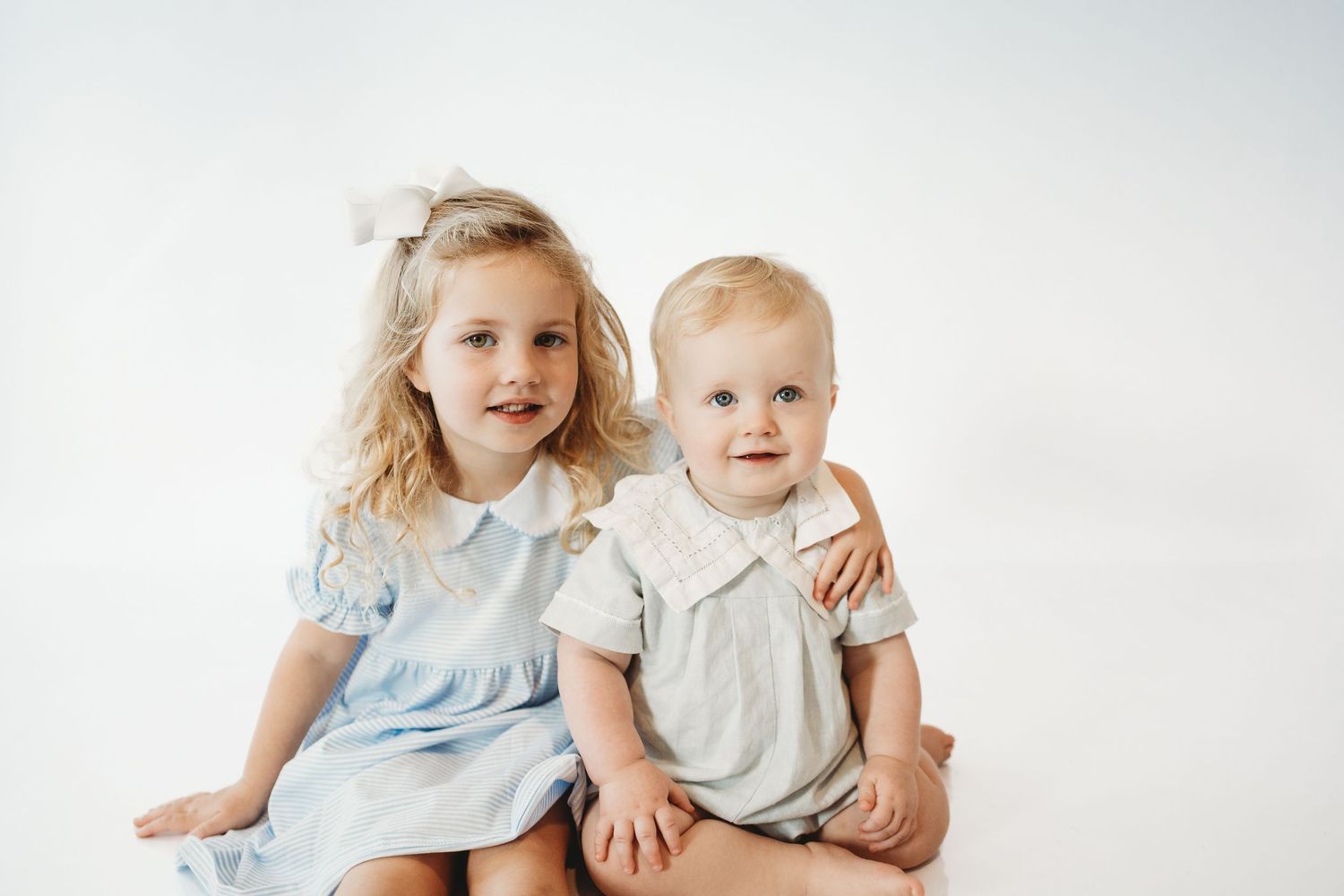Two young children in light-colored outfits sitting together against a white background.