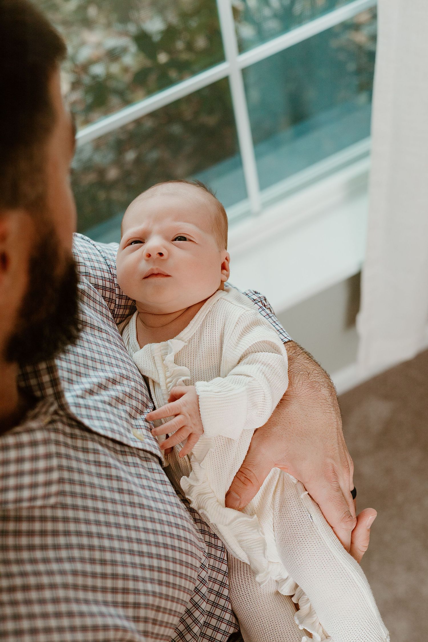 A newborn baby wearing white sleeps peacefully while being cradled by someone in a plaid shirt.
