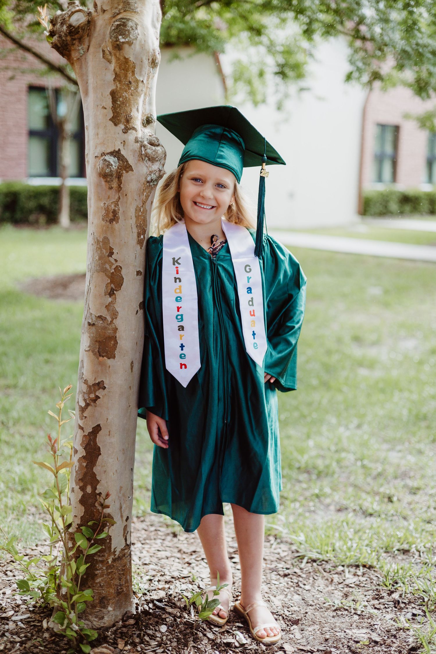 A graduate in a green cap and gown poses near a tree trunk during an outdoor photo session.