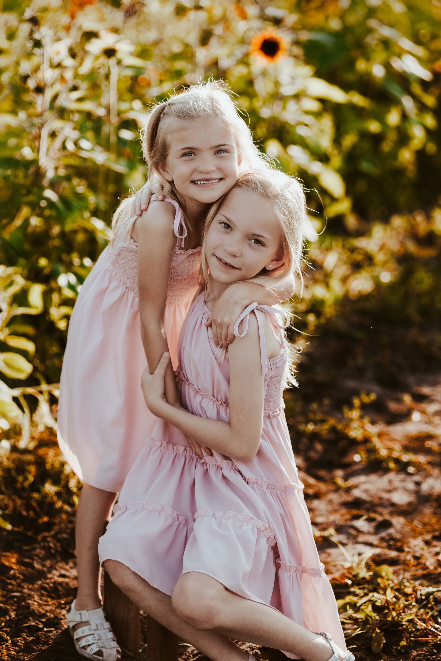 Two children in pink dresses embrace among sunflowers during a sunny outdoor photoshoot.