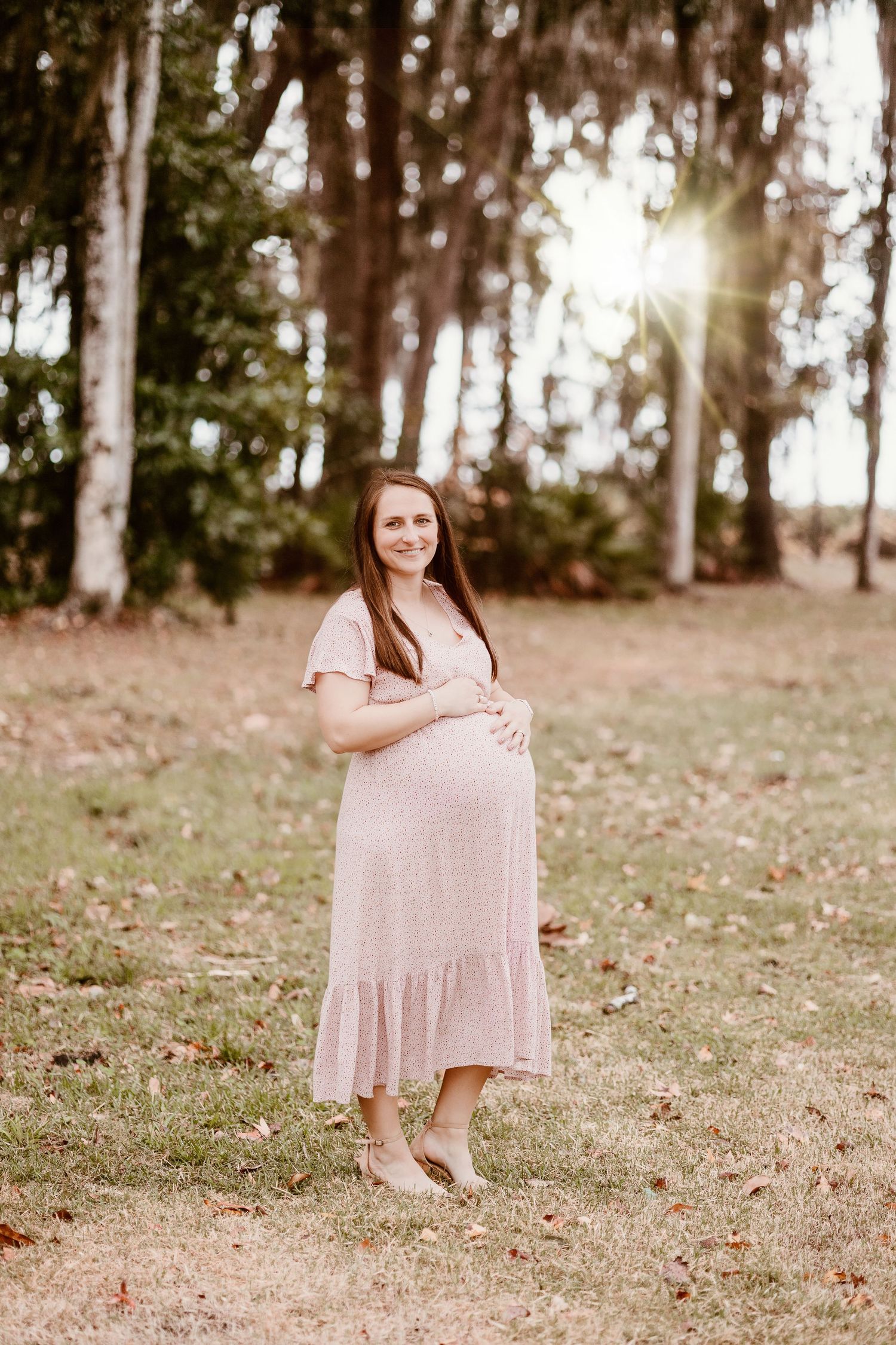 Maternity photo session in a wooded setting with woman in flowing blush dress.