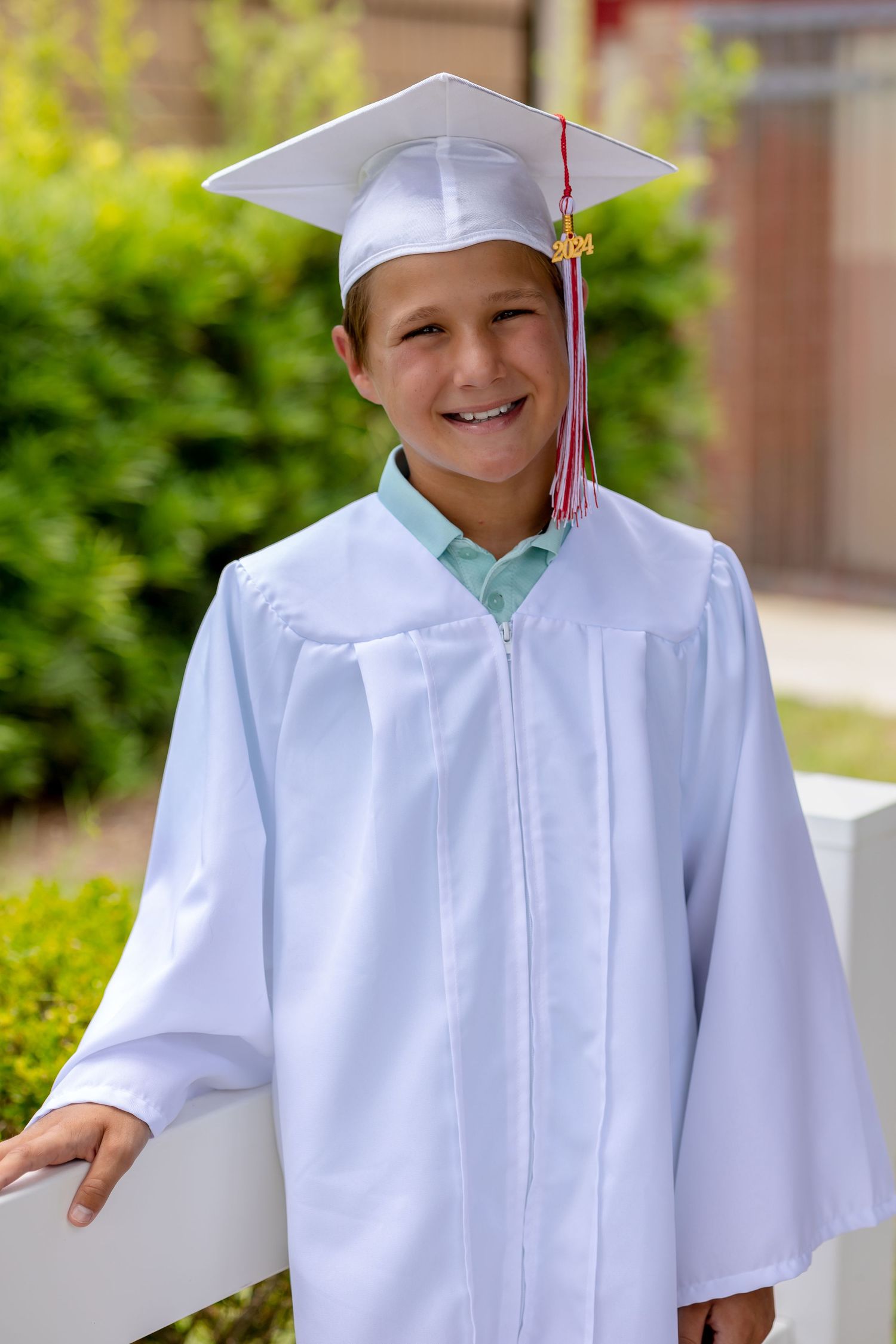 Young graduate in white cap and gown smiling outdoors during kindergarten or elementary school graduation ceremony.