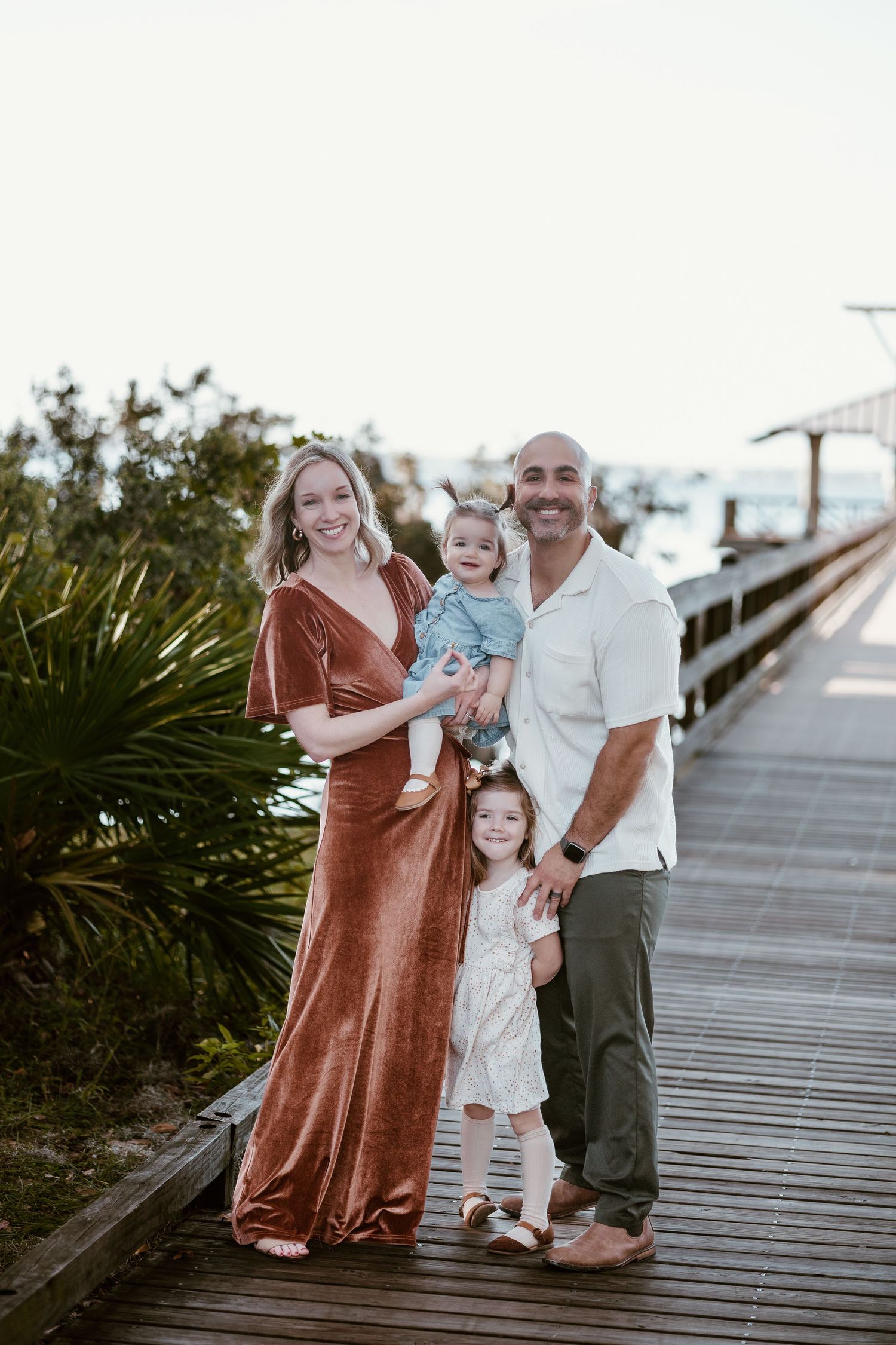 A family poses together on a wooden boardwalk with coastal vegetation in the background.