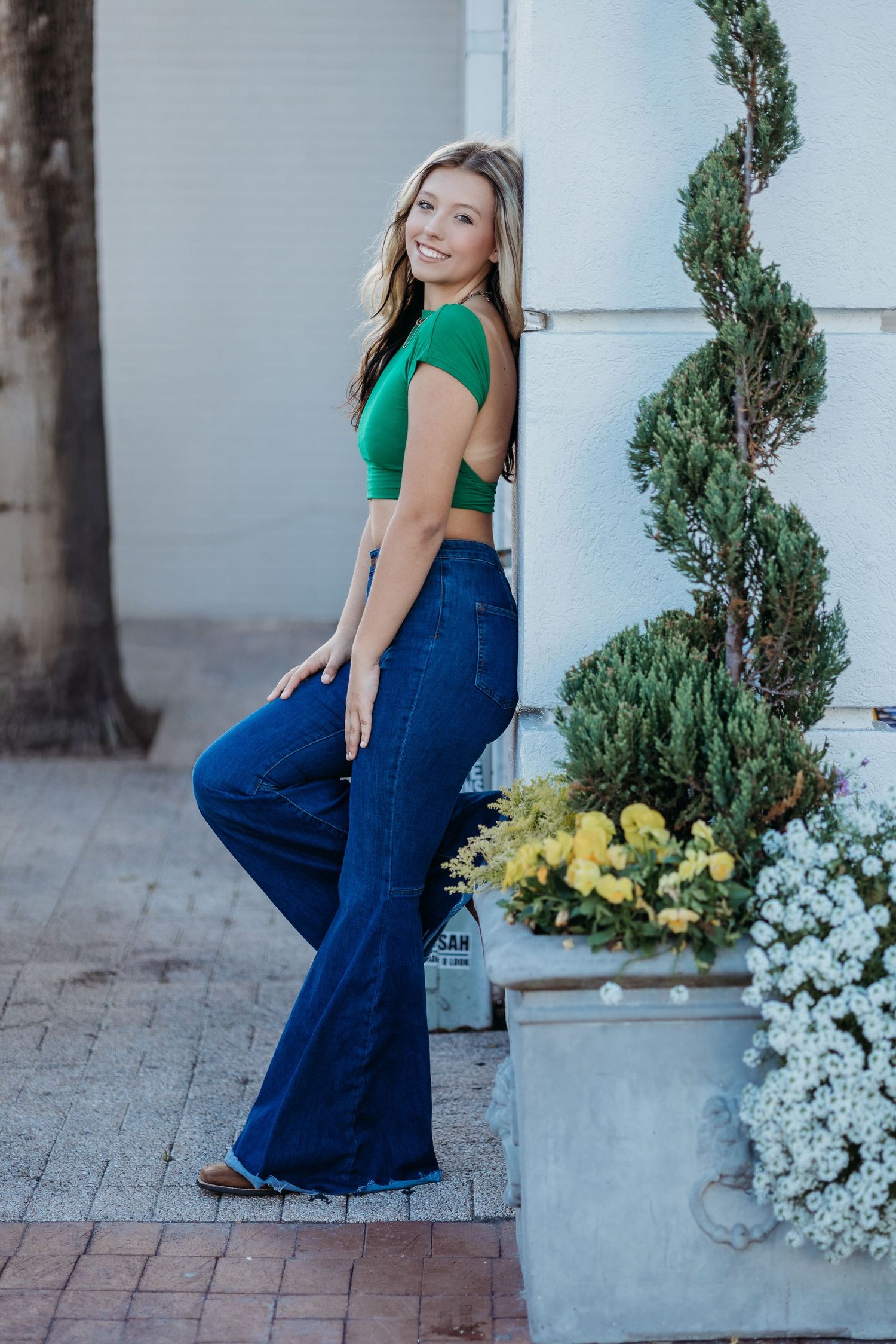 Fashion poses against a white wall with decorative plants wearing green top and flared jeans.