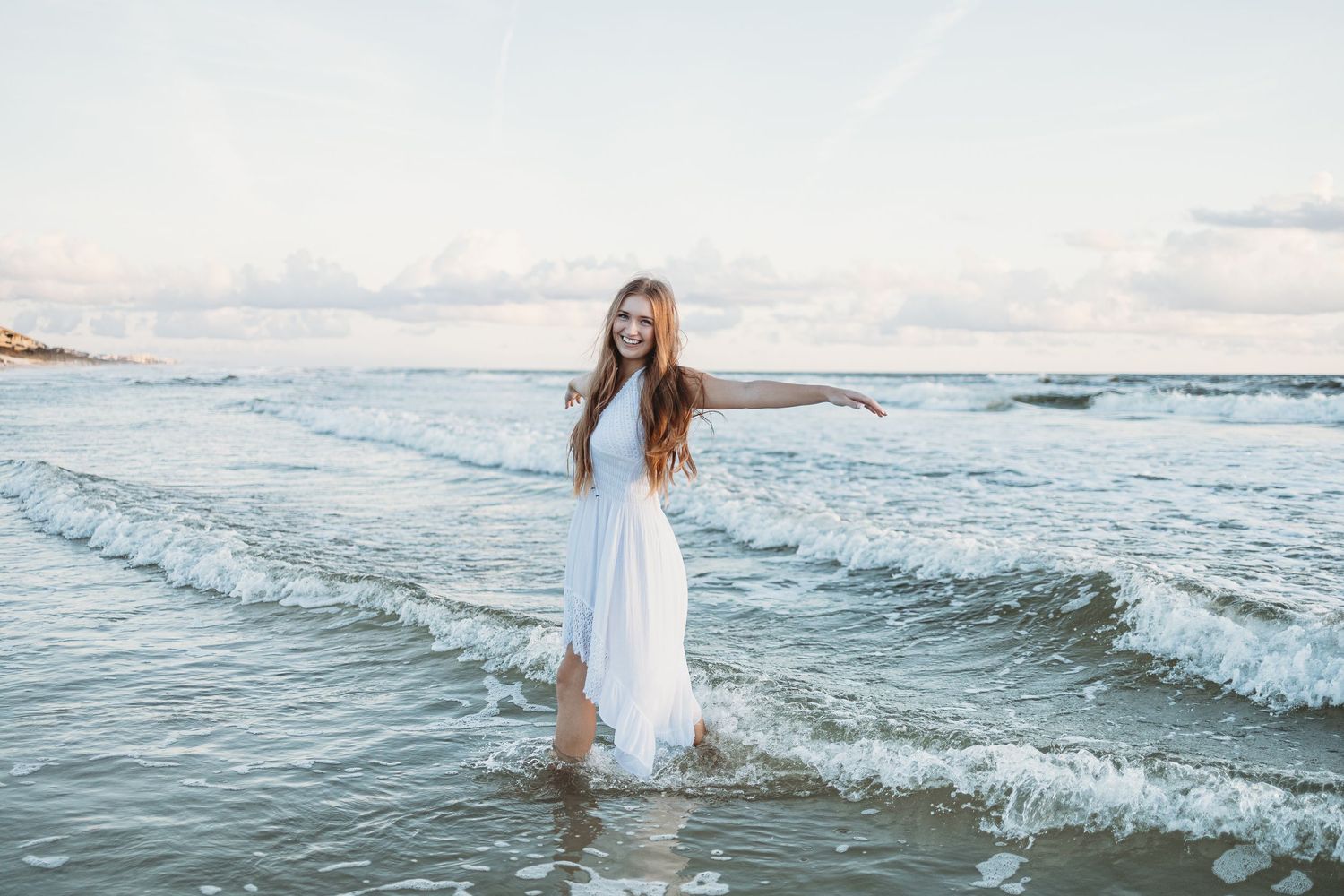 A person in a flowing white dress walks along the shoreline as ocean waves roll in during sunset.