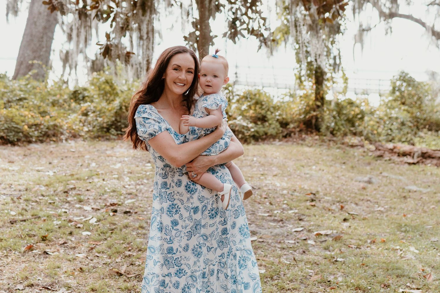 Woman in floral blue dress holds baby while standing in outdoor garden setting with Spanish moss trees.