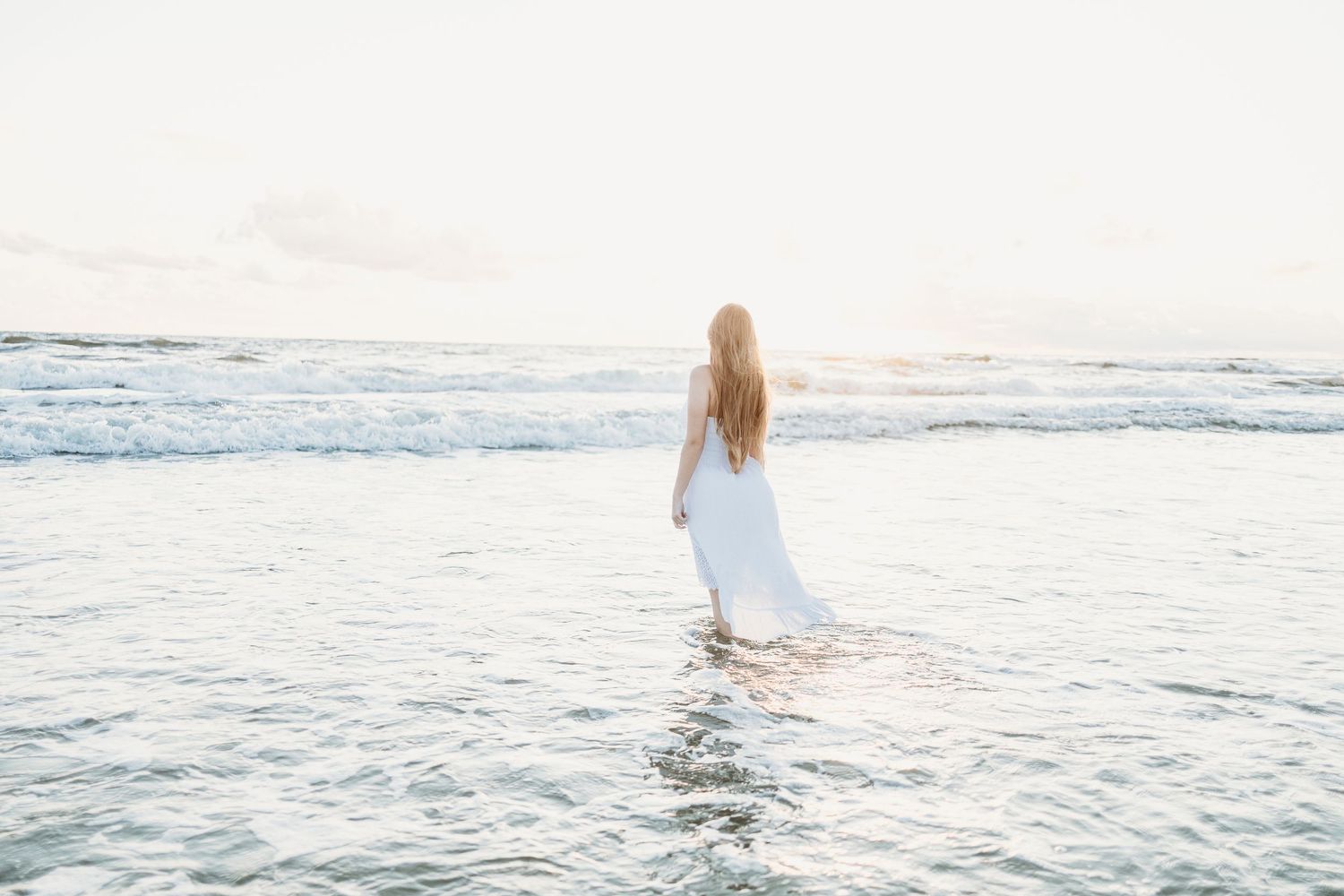 A woman in a white dress wades into the ocean waves at sunset, creating a dreamy coastal scene.