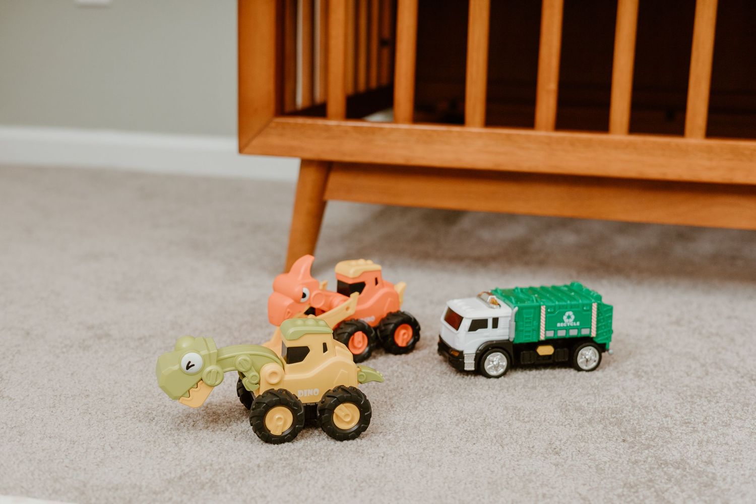 Colorful toy vehicles and trucks are displayed on a carpeted floor near wooden furniture.