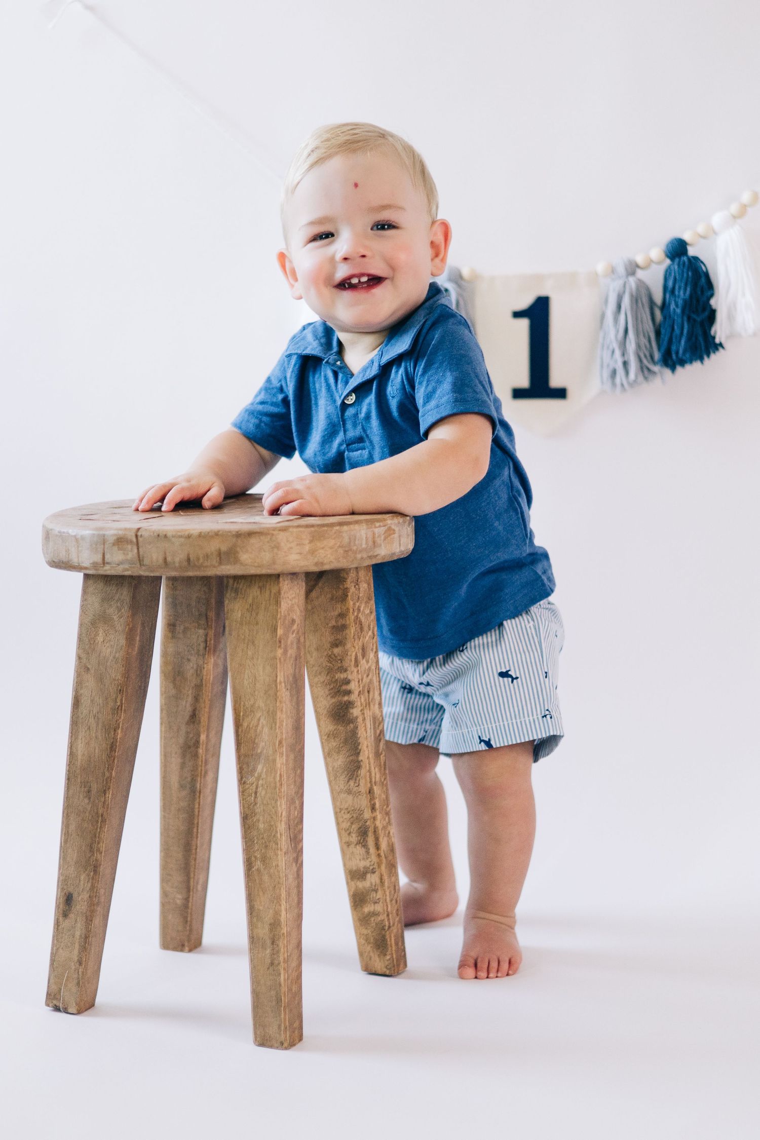 Smiling baby in blue shirt leaning on wooden stool with party decorations.