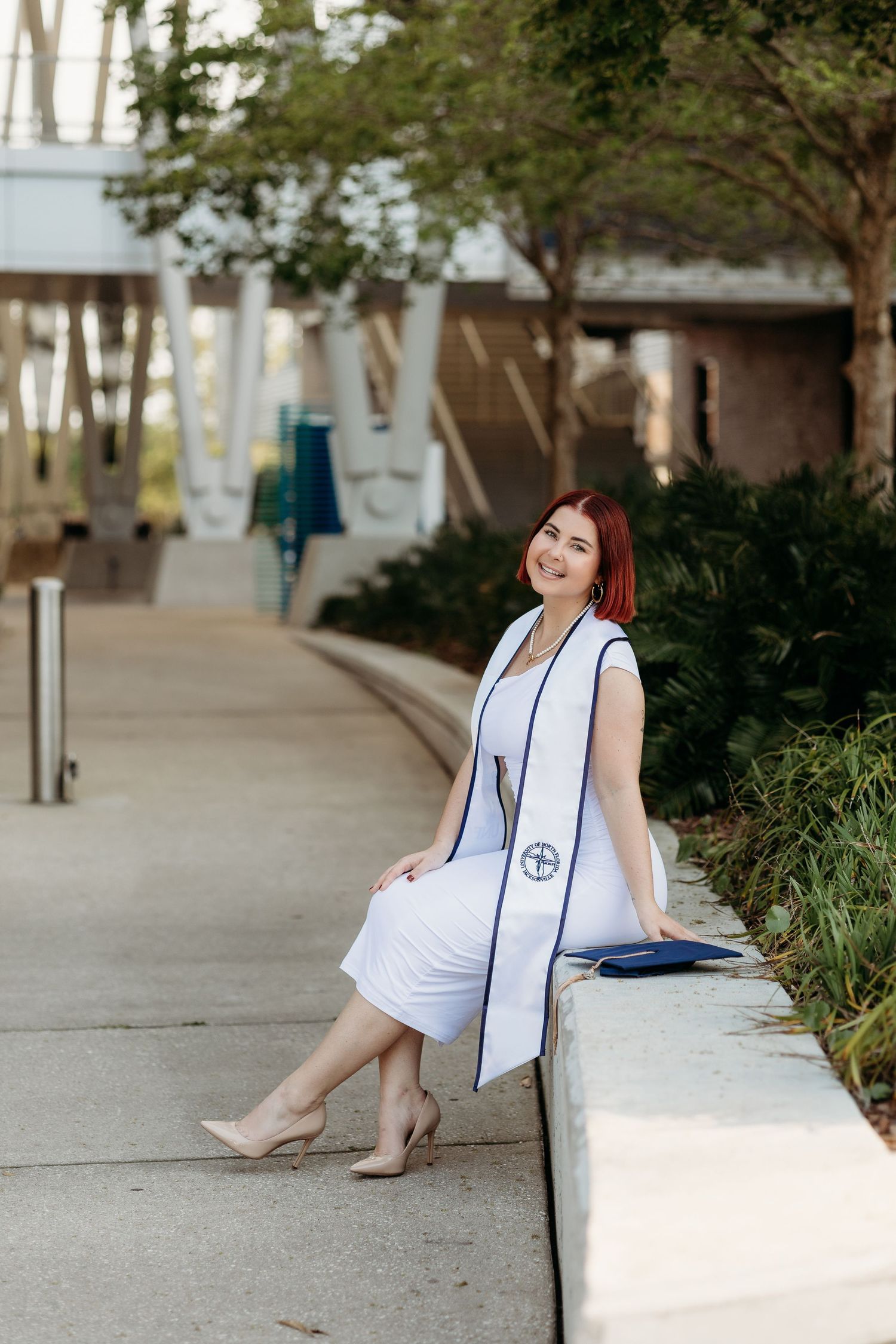 Graduate in white stole sitting on concrete ledge for outdoor portraits.