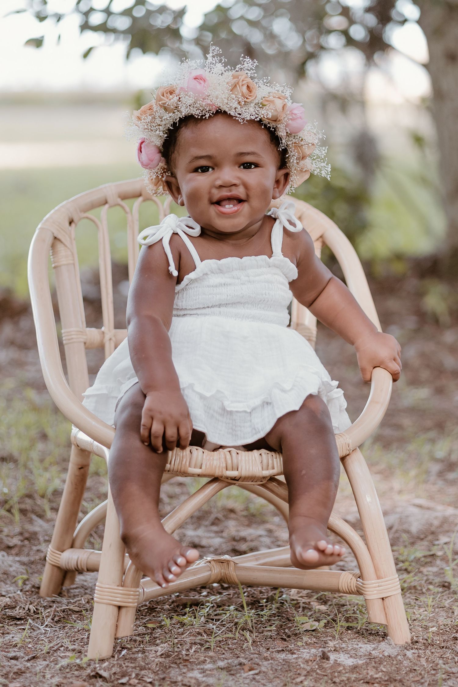 Small child sits in wooden chair outdoors wearing white outfit and floral crown.