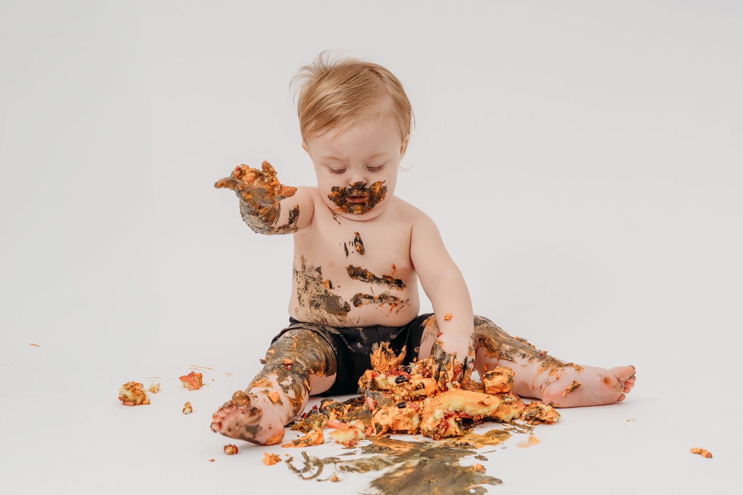 Baby wearing white outfit sits on floor playing with cake and frosting during cake smash photo session.