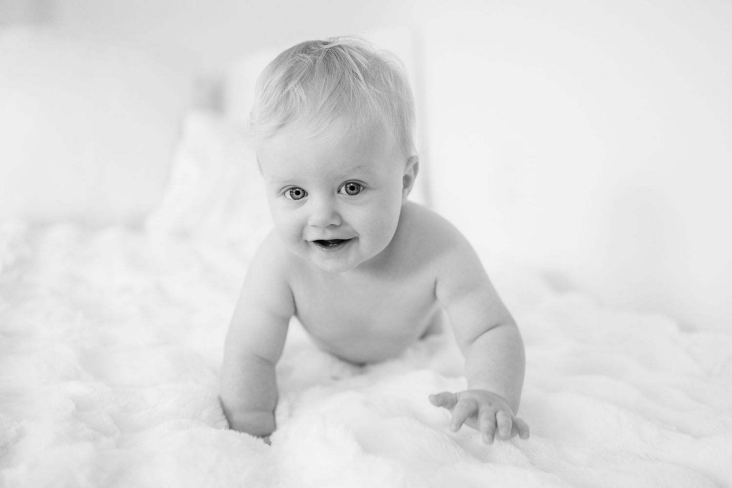 A black and white portrait of a baby lying on soft white blankets with a joyful expression.