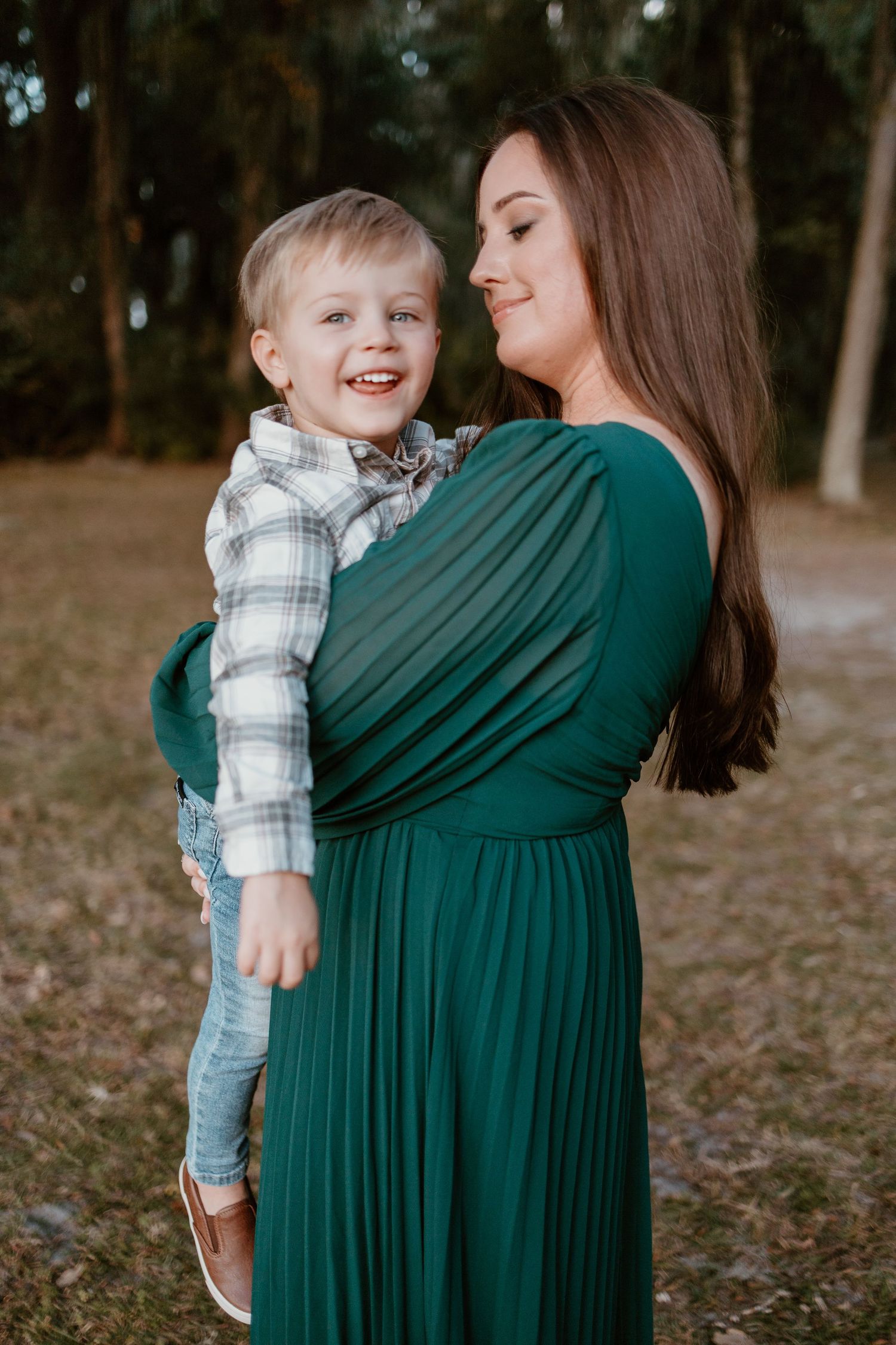 A sweet moment between a child in plaid and a person wearing an emerald green dress in a wooded outdoor setting.