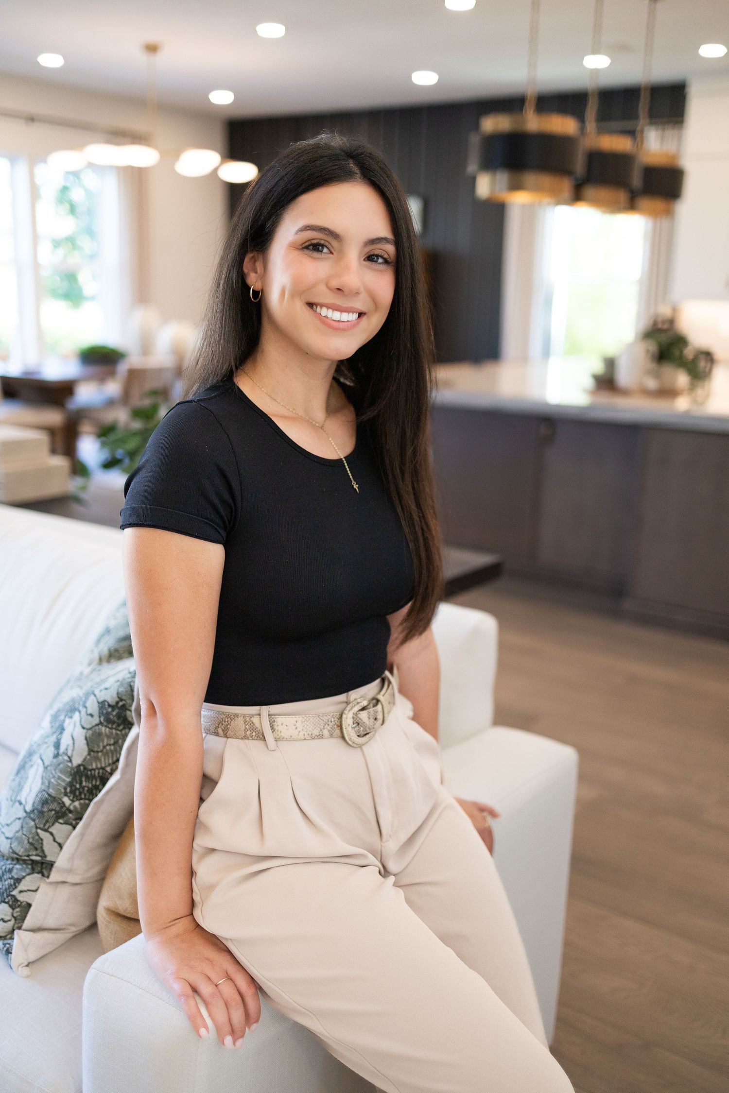 Woman in black t-shirt and beige pants sitting casually on modern white couch in bright contemporary living space.