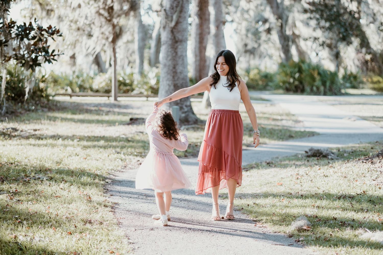 A mother and daughter walk together on a sunlit path through a dreamy outdoor setting.