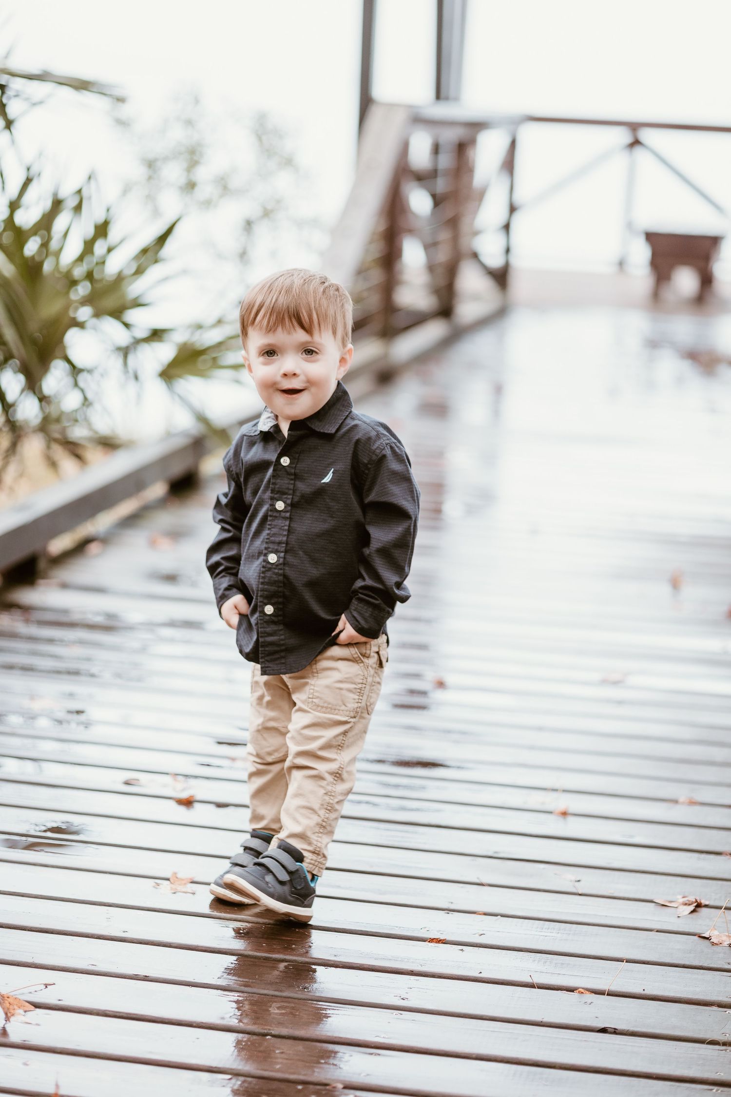 Toddler in black button-up shirt and khakis walking on wet wooden deck.