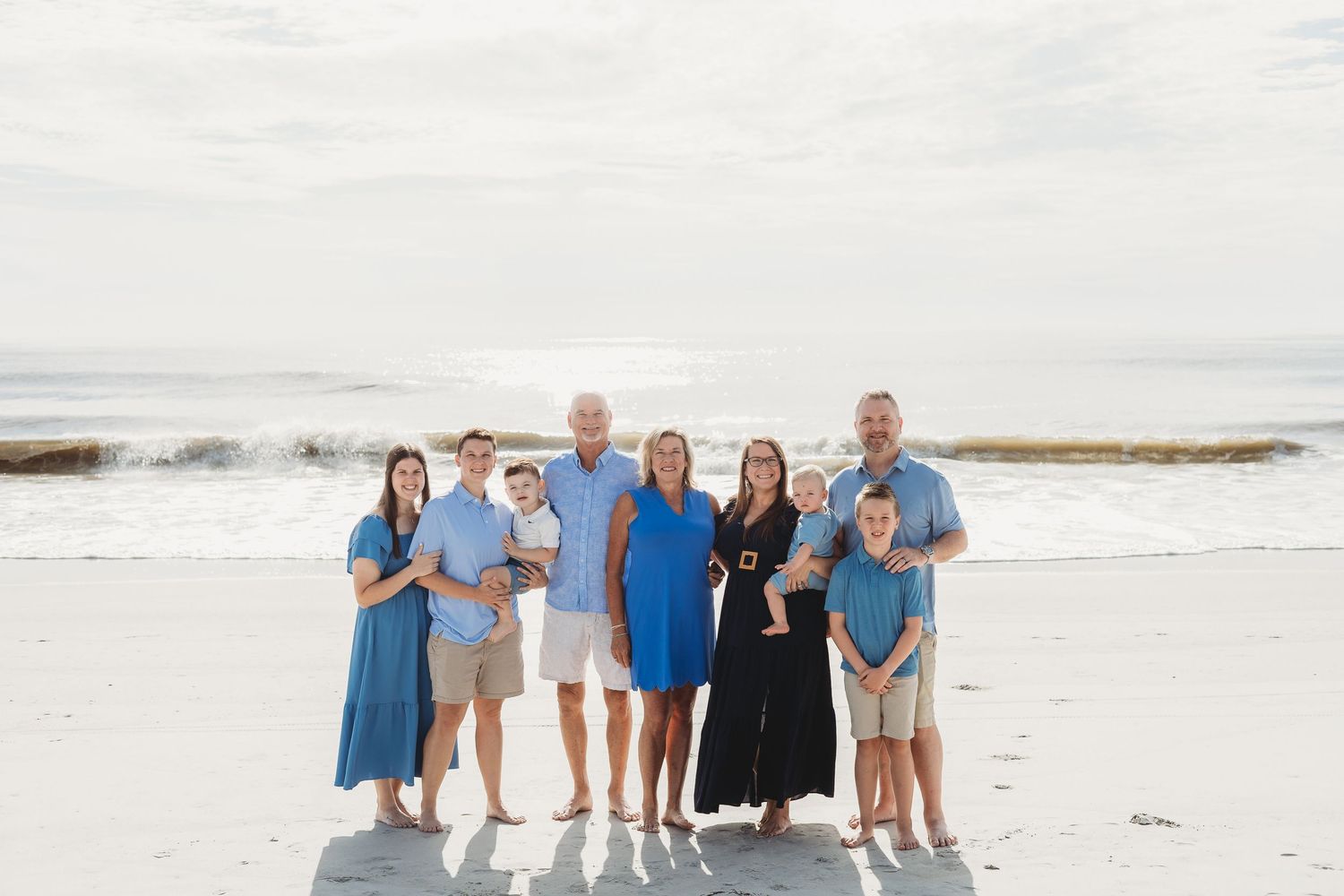 A large family group poses together on the beach wearing coordinated blue and white outfits.