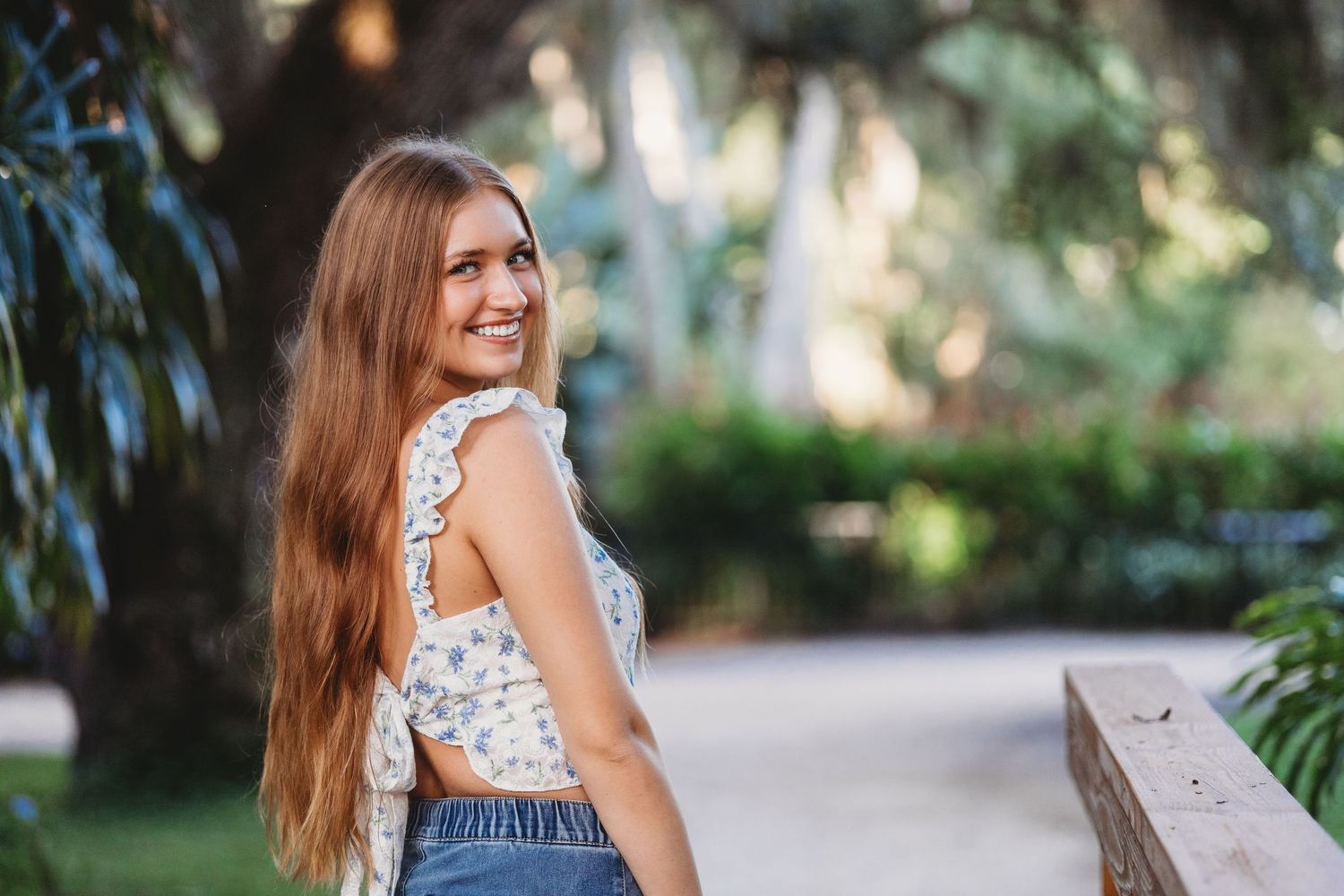 Woman in floral dress poses outdoors on a garden bench during a sunny summer day.