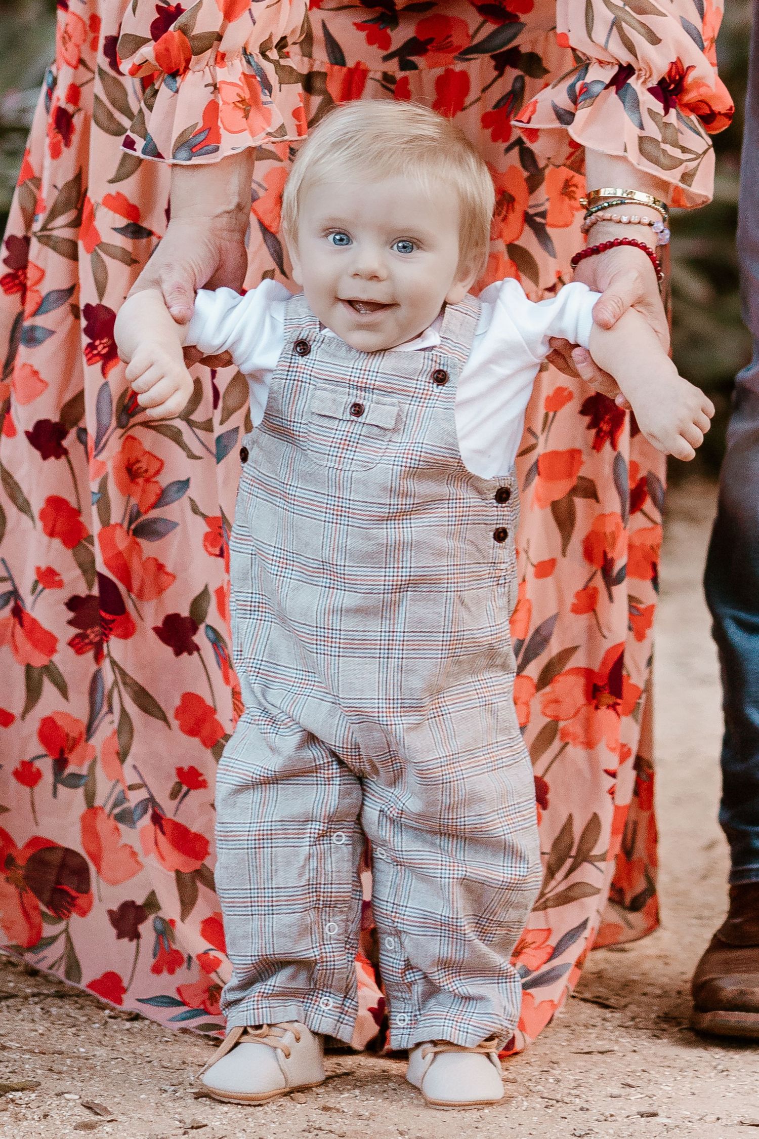Baby in white overalls stands against a coral floral backdrop in a series of sweet milestone photos.