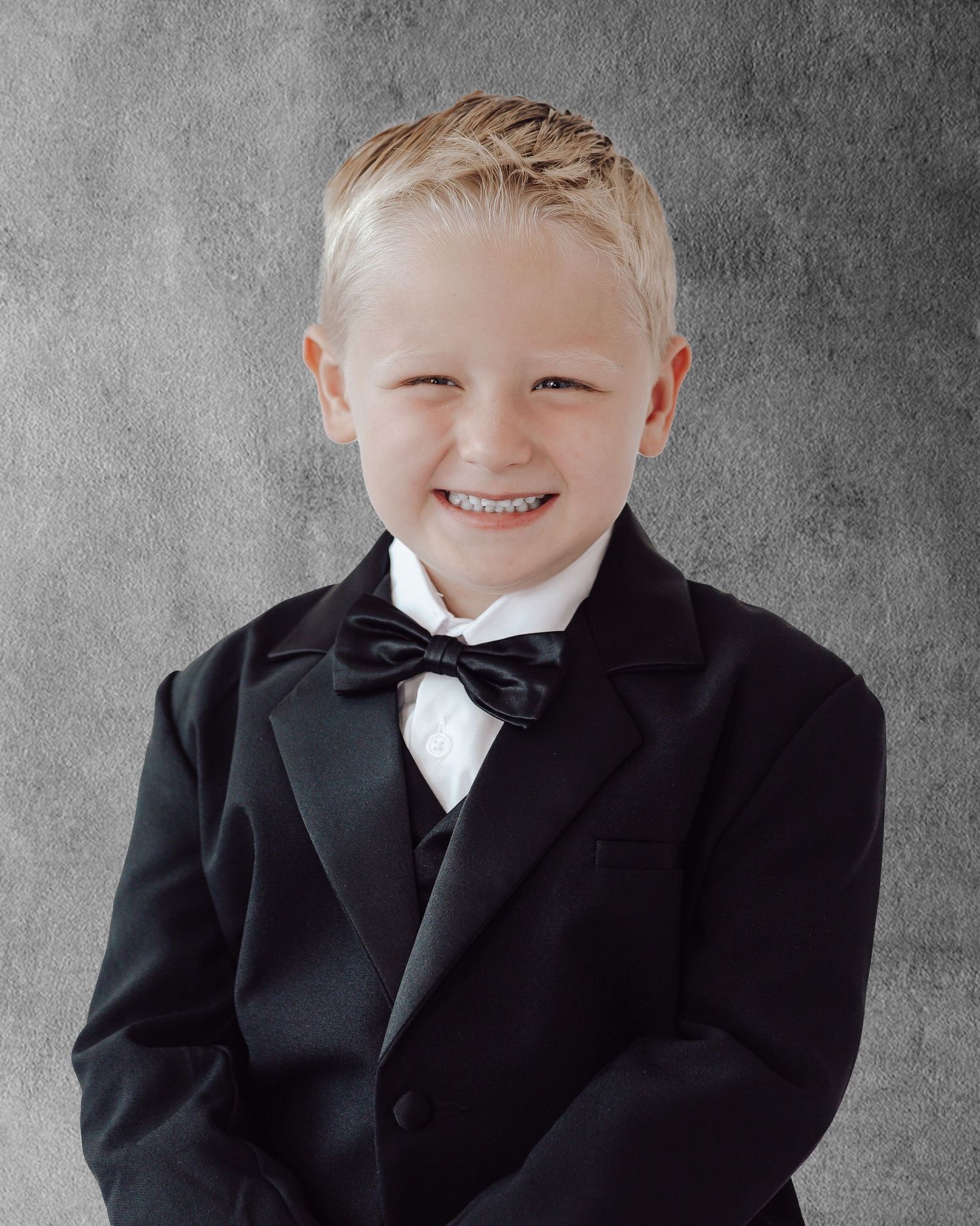 Child in black tuxedo and bowtie smiling in professional studio photoshoot.