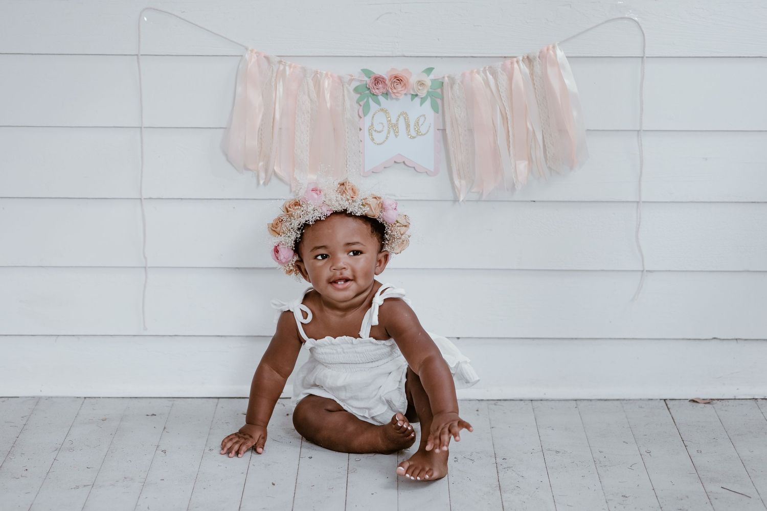 Baby in white outfit sits on wooden chair against white wall backdrop.