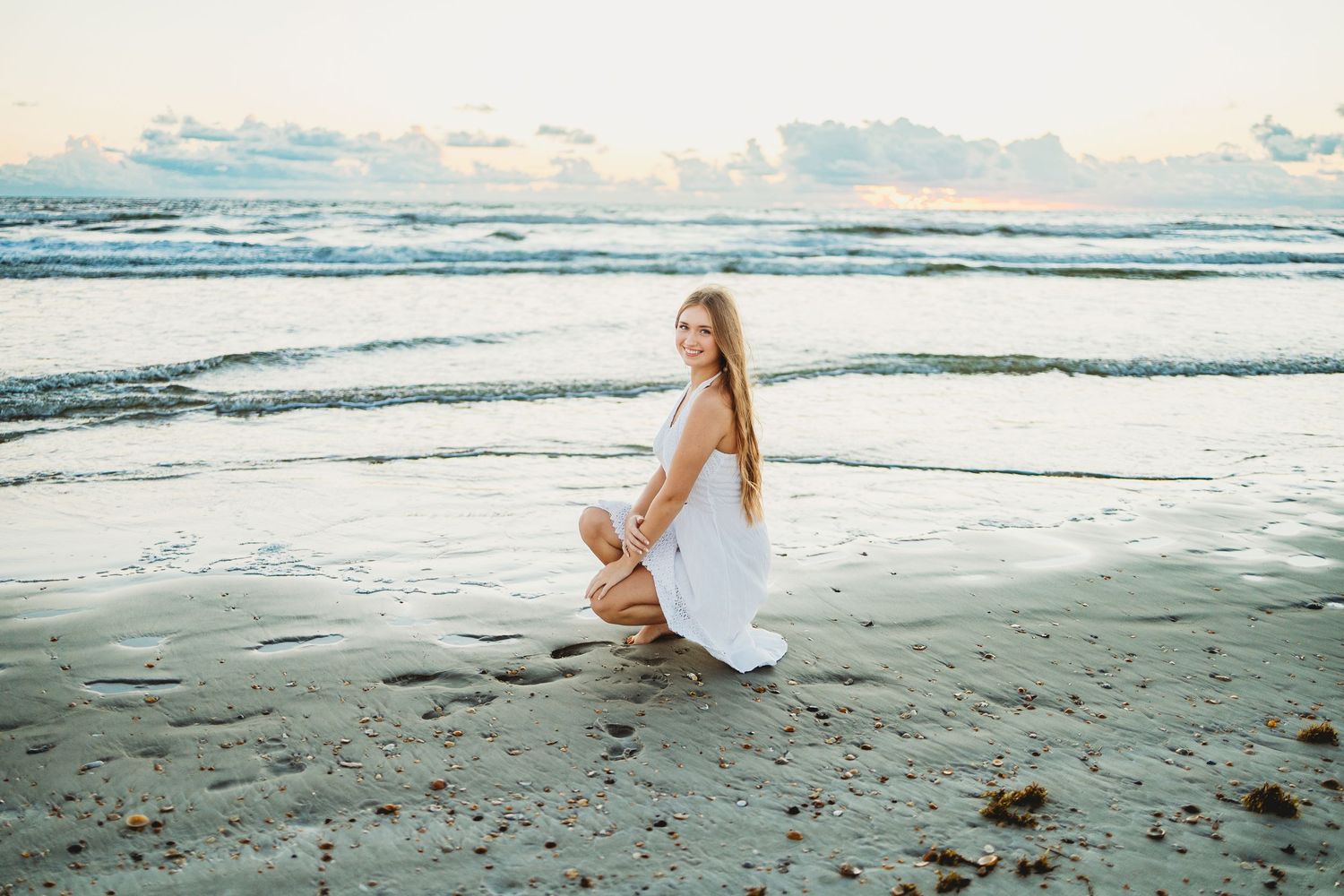 A person in a white dress sits peacefully on a sandy beach at sunset while waves roll in from the ocean.