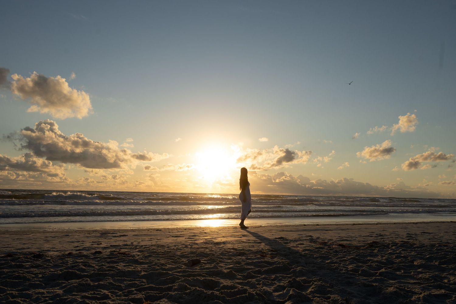Silhouetted figure walking along a beach at sunset with dramatic clouds and golden light reflecting on wet sand.