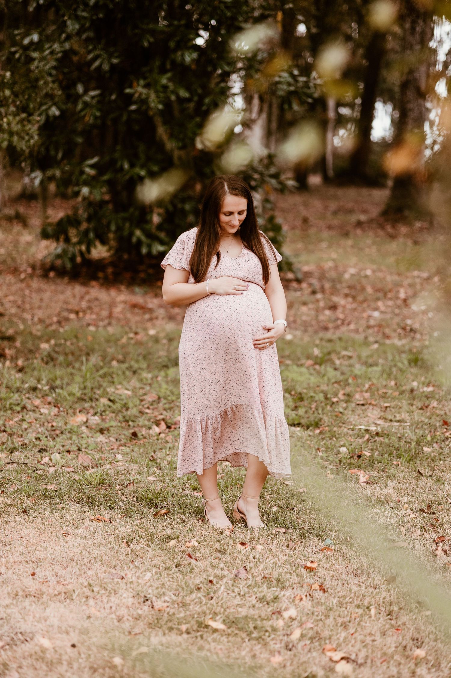 Expectant mother in pink dress poses gracefully in natural outdoor setting.