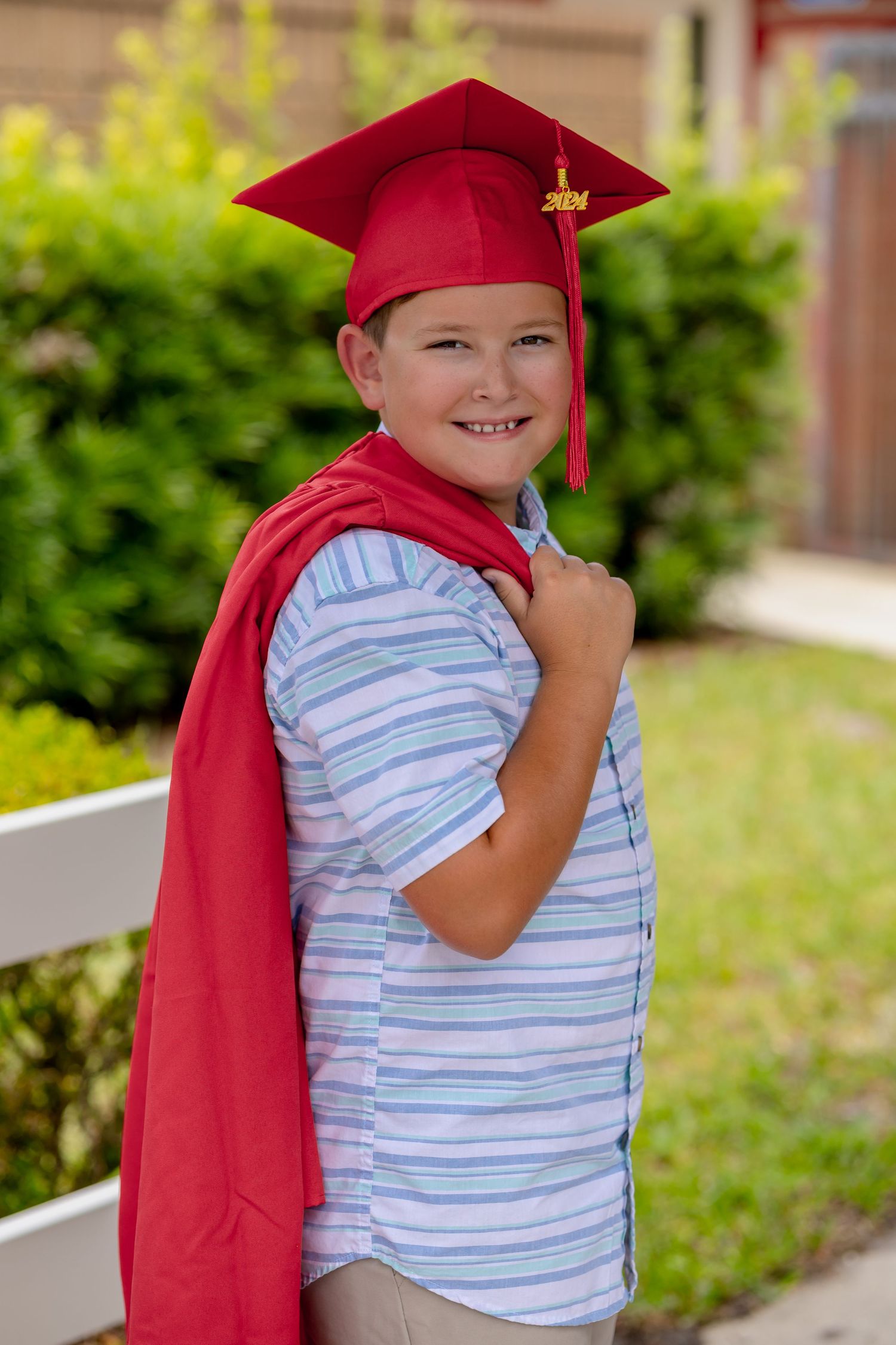 A young student in a red graduation cap and striped shirt smiles while standing outdoors.