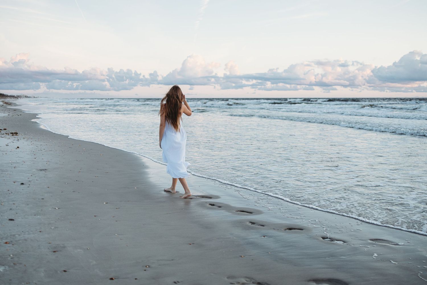 Person in a white dress walks barefoot along a serene beach shoreline at dusk with waves and mountains in the distance.