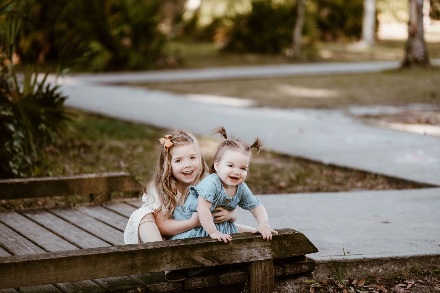 Two small children playfully lean over a wooden boardwalk in matching white outfits.