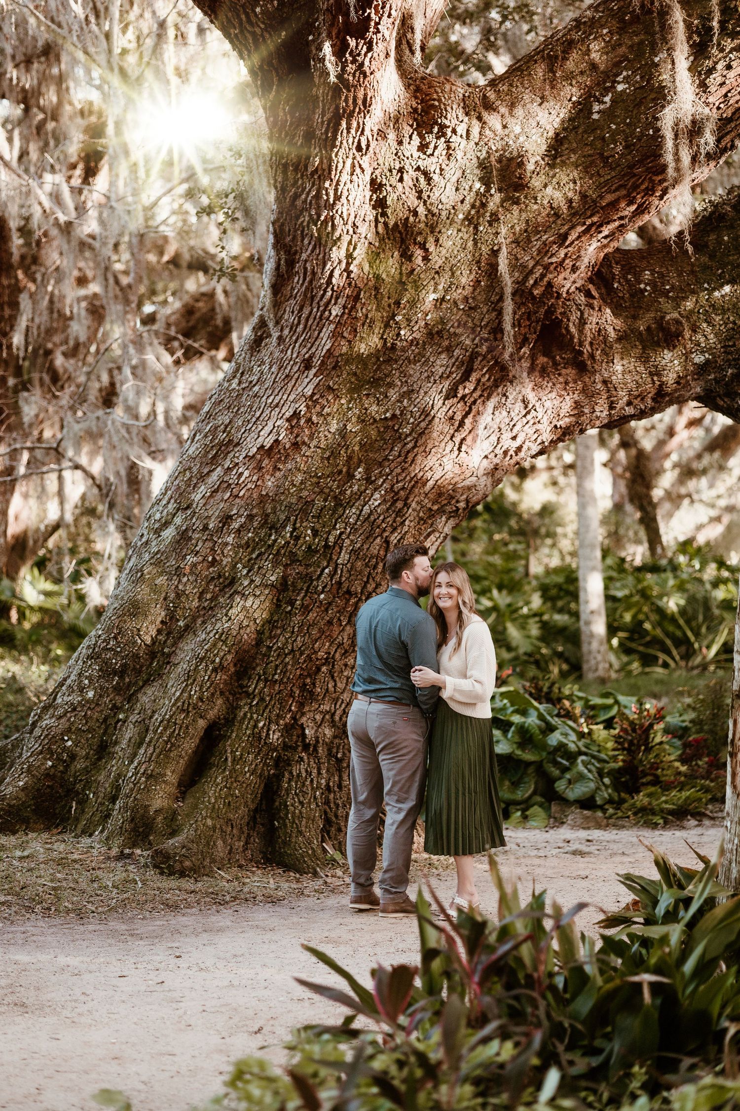 A romantic moment captured under a majestic old tree in a serene outdoor setting.