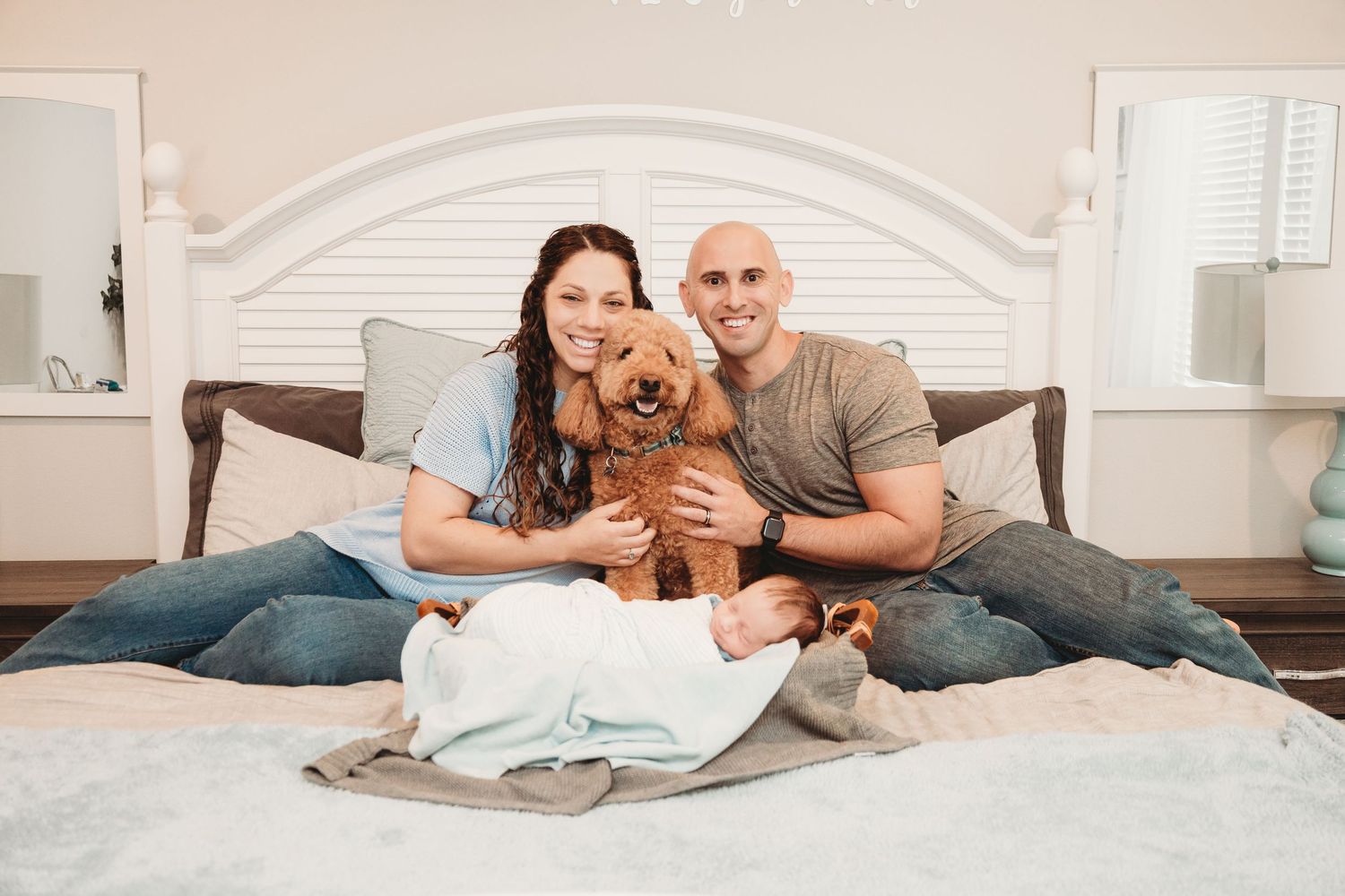 Family lounging together on bed with their pet dog and newborn.