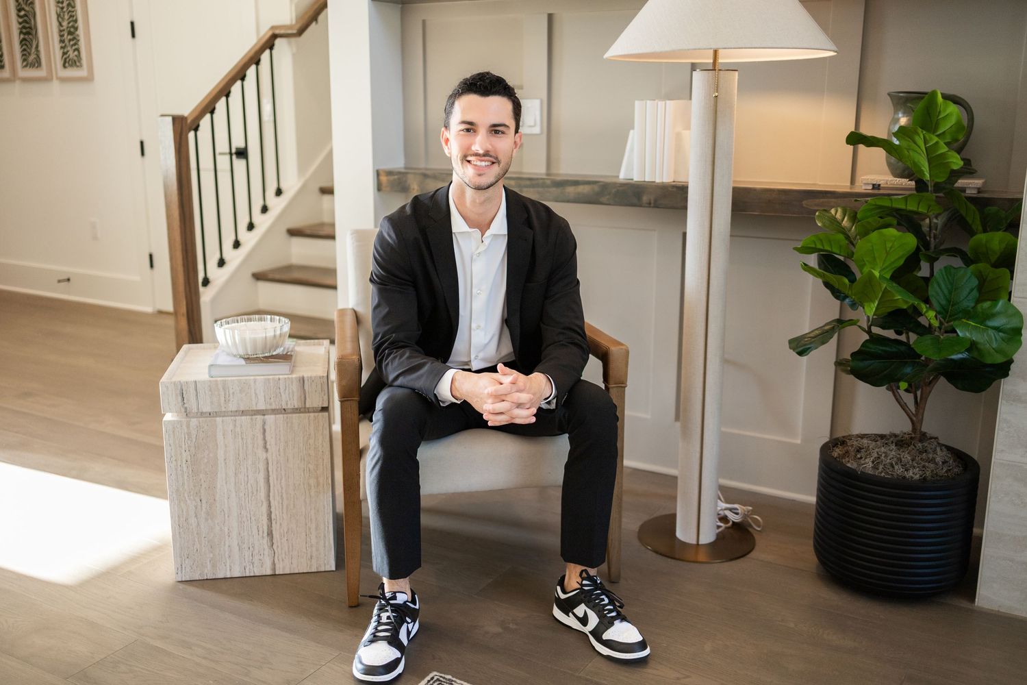 Professional wearing a dark suit and sneakers sits casually in a modern minimalist interior with white walls and plants.