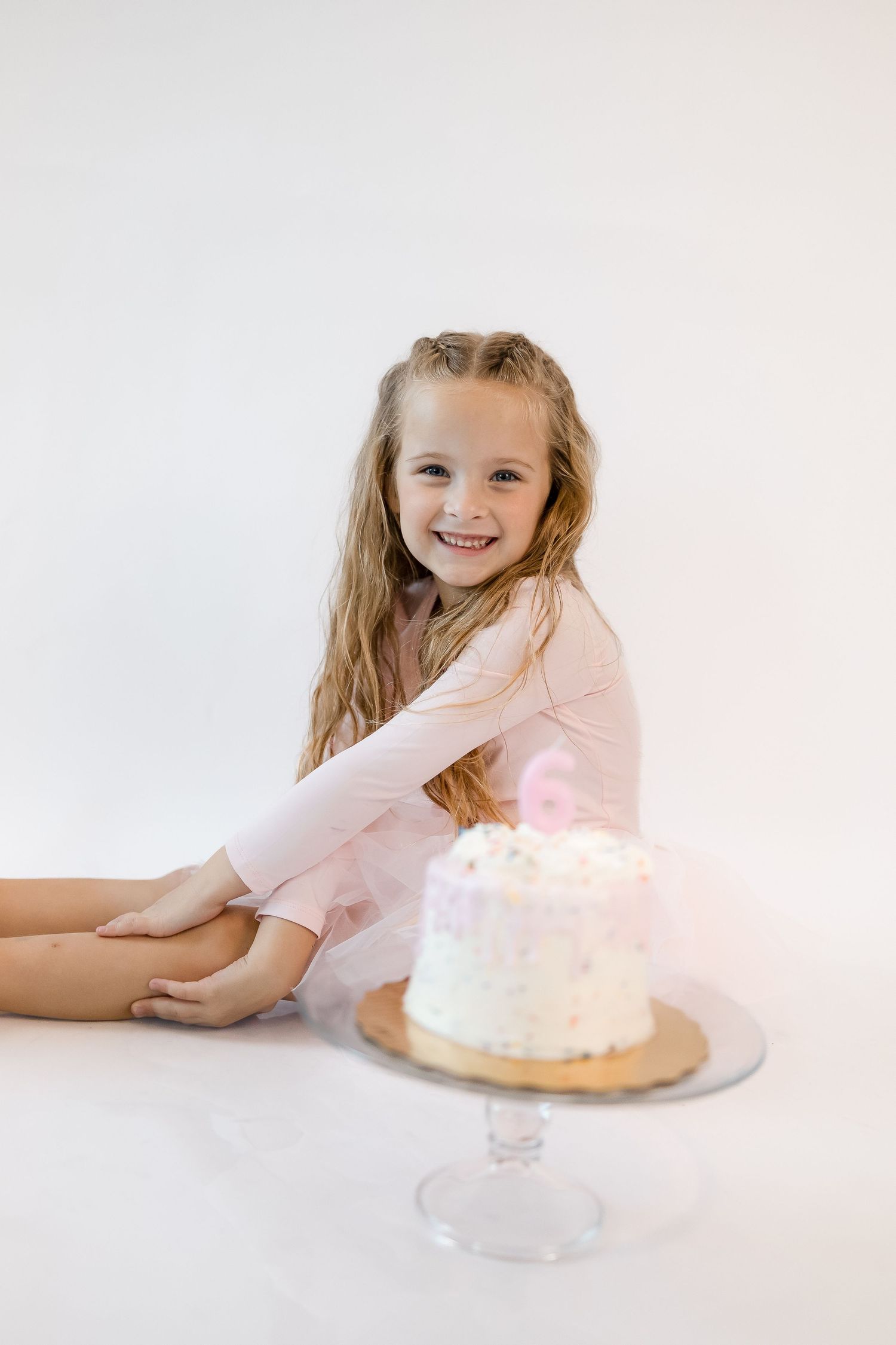 A child in pink dress poses playfully with a white frosted birthday cake on a light background.