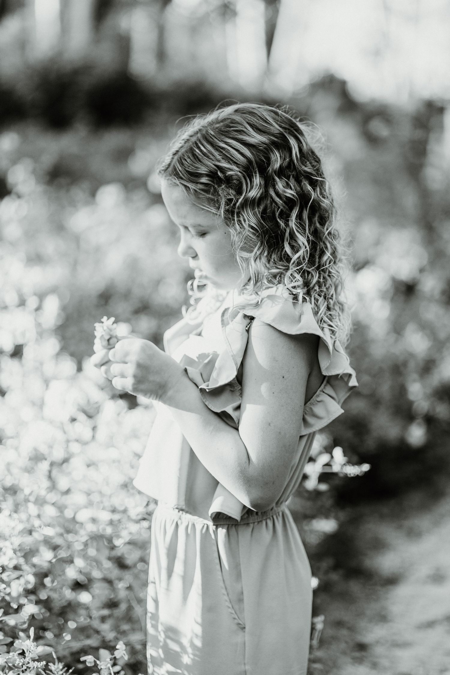 Black and white portrait of a young child with curly hair in a ruffled dress examining flowers in a garden.