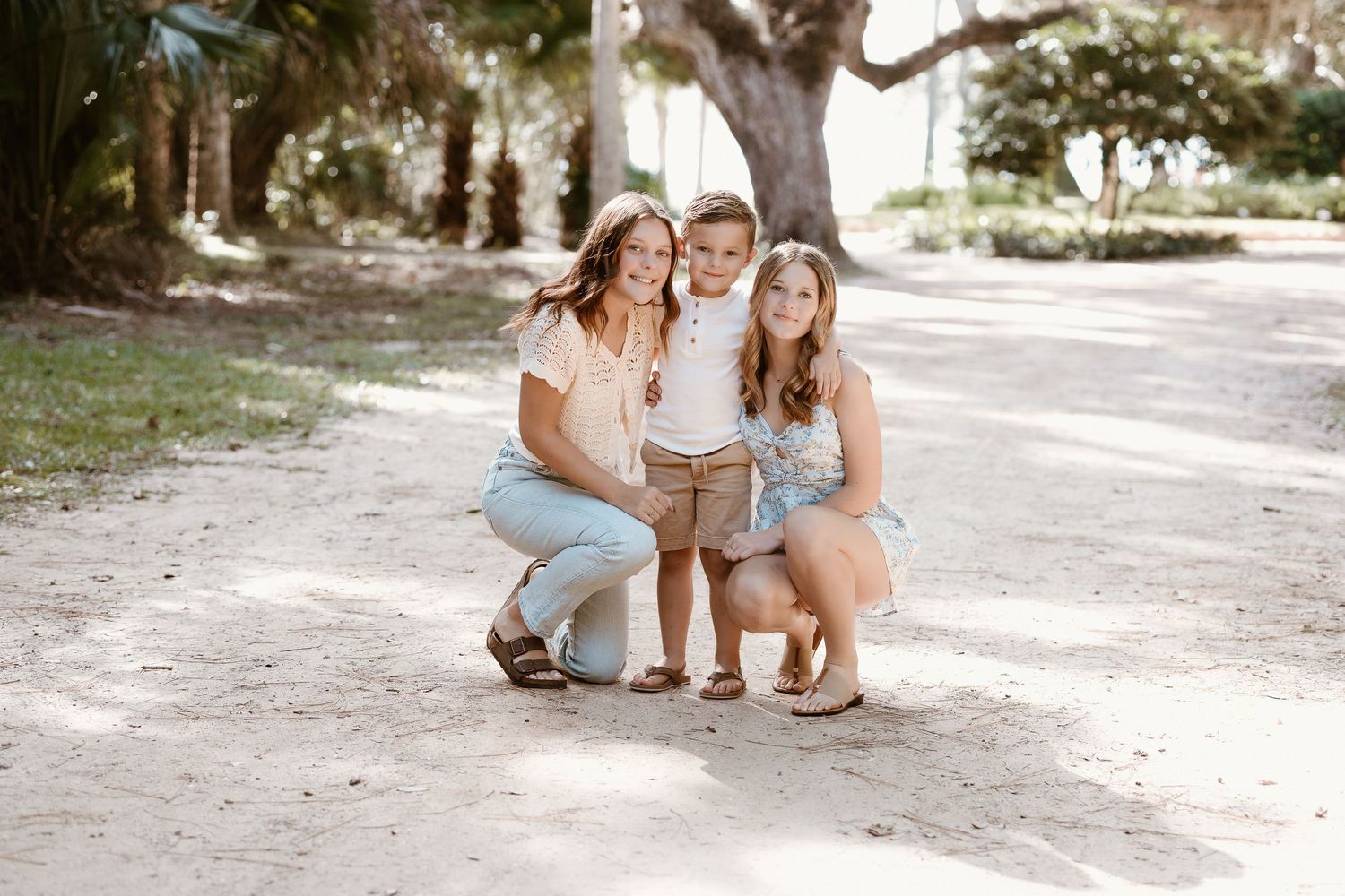 Three people crouching together on a sunlit dirt path surrounded by trees in a natural outdoor setting.