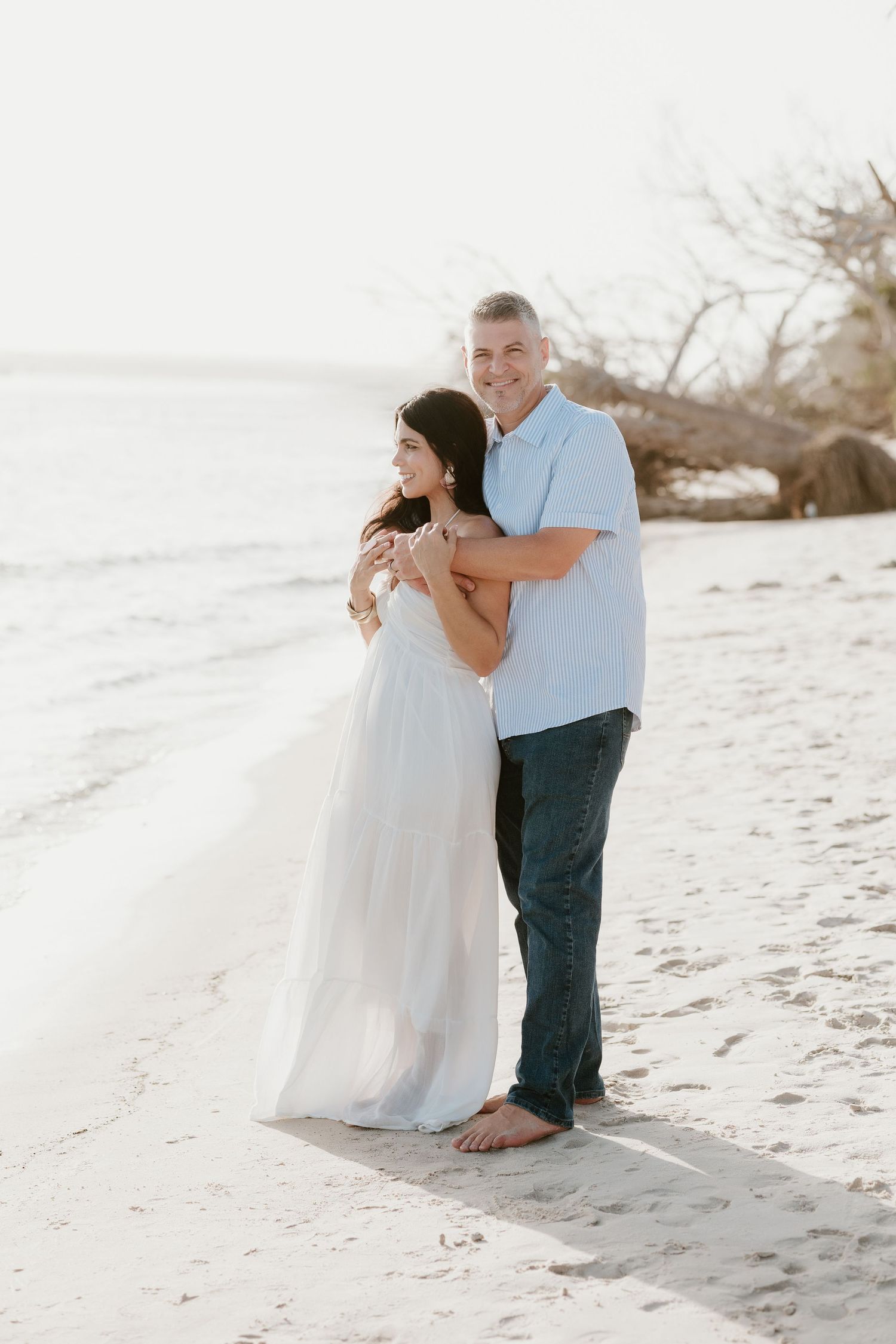 A couple embraces romantically on a sandy white beach during a photoshoot.