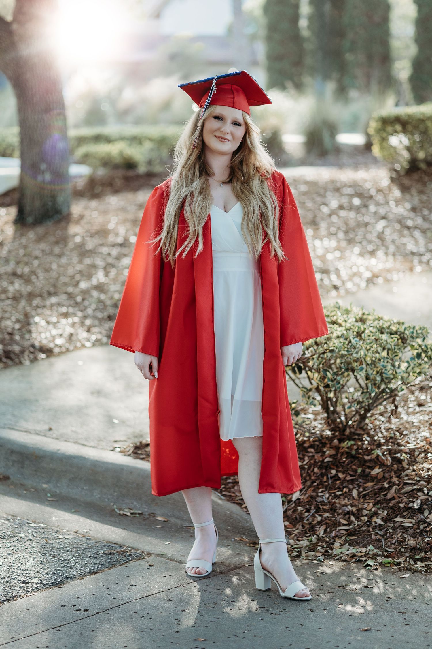 Graduate in red cap and gown walking outdoors on concrete path.