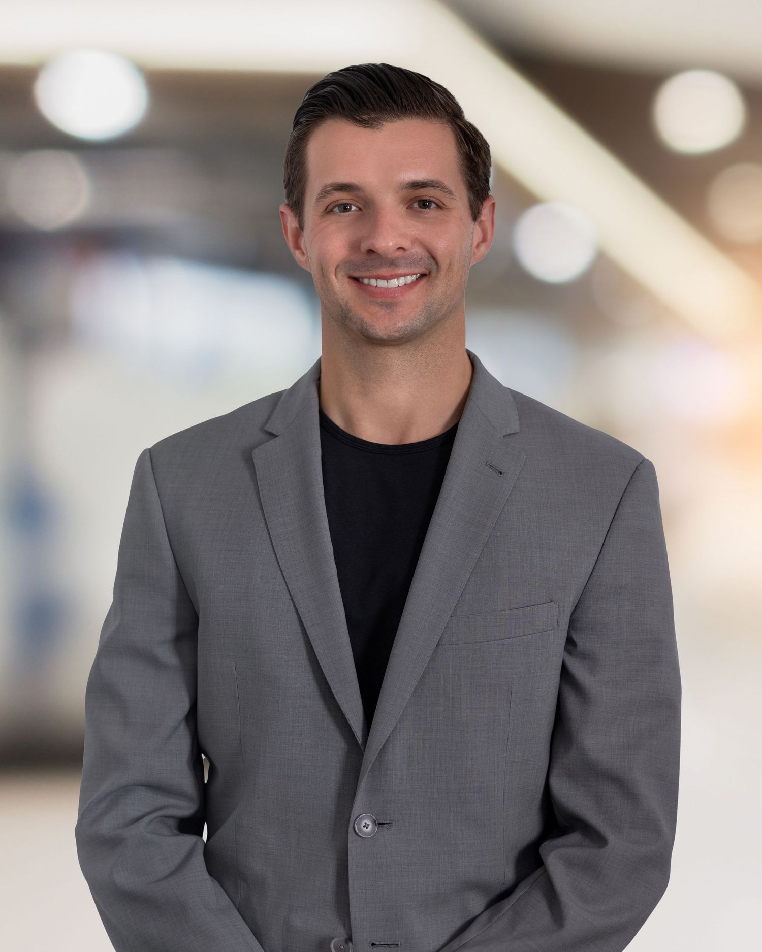 Professional headshots of person in grey suit and black shirt against bright background.
