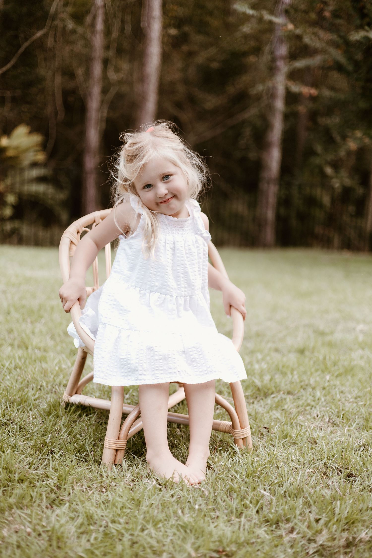 Small child in white dress sits playfully on wooden chair in grassy outdoor setting.