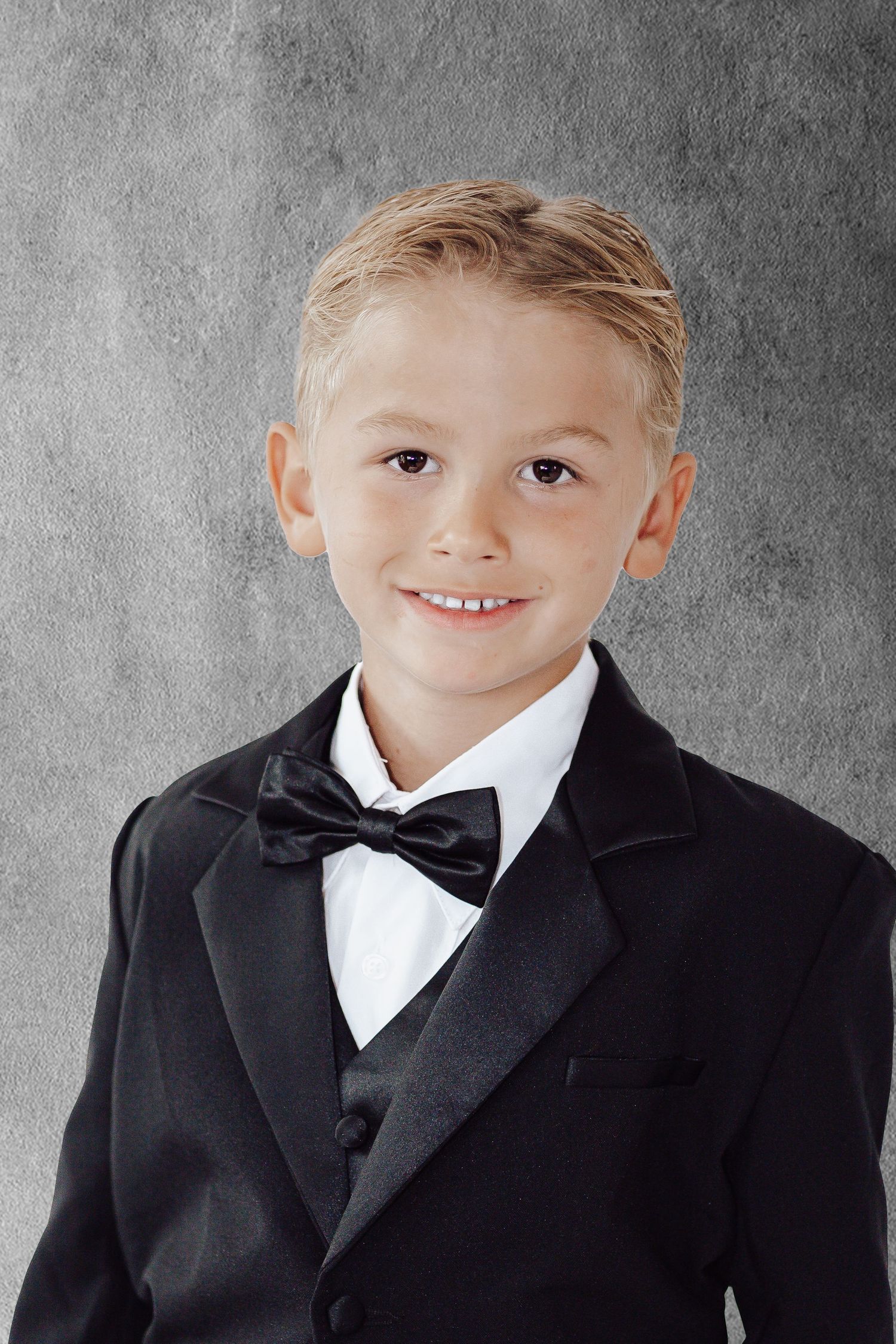 Studio portraits of child wearing black formal suit with white bowtie against grey backdrop.