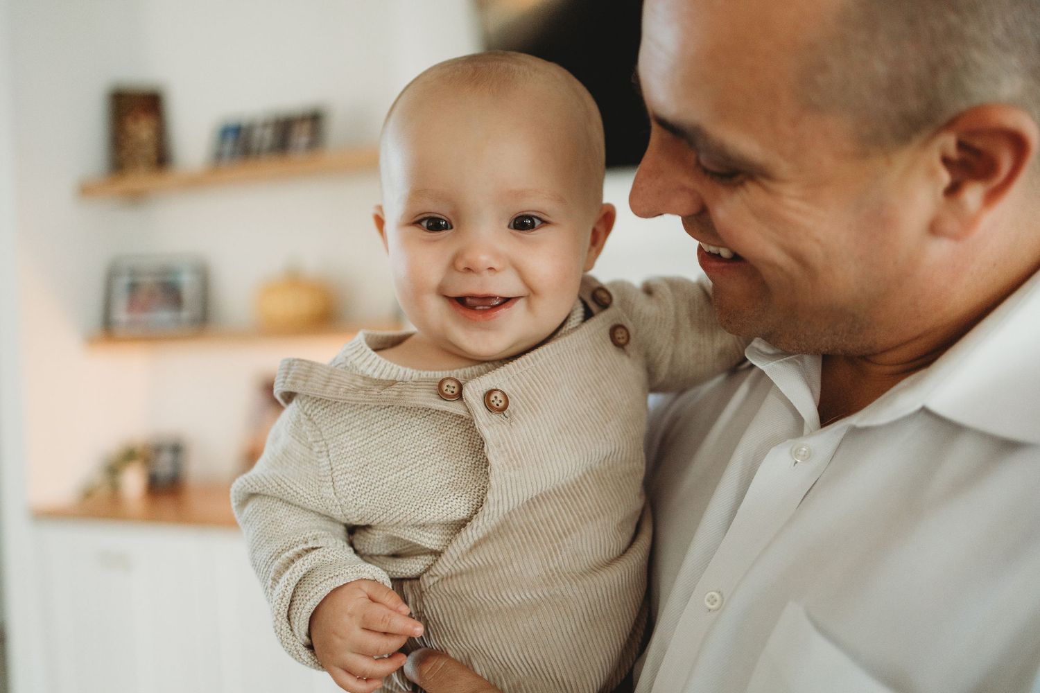 Baby in beige outfit smiling and playing while being held in loving embrace.