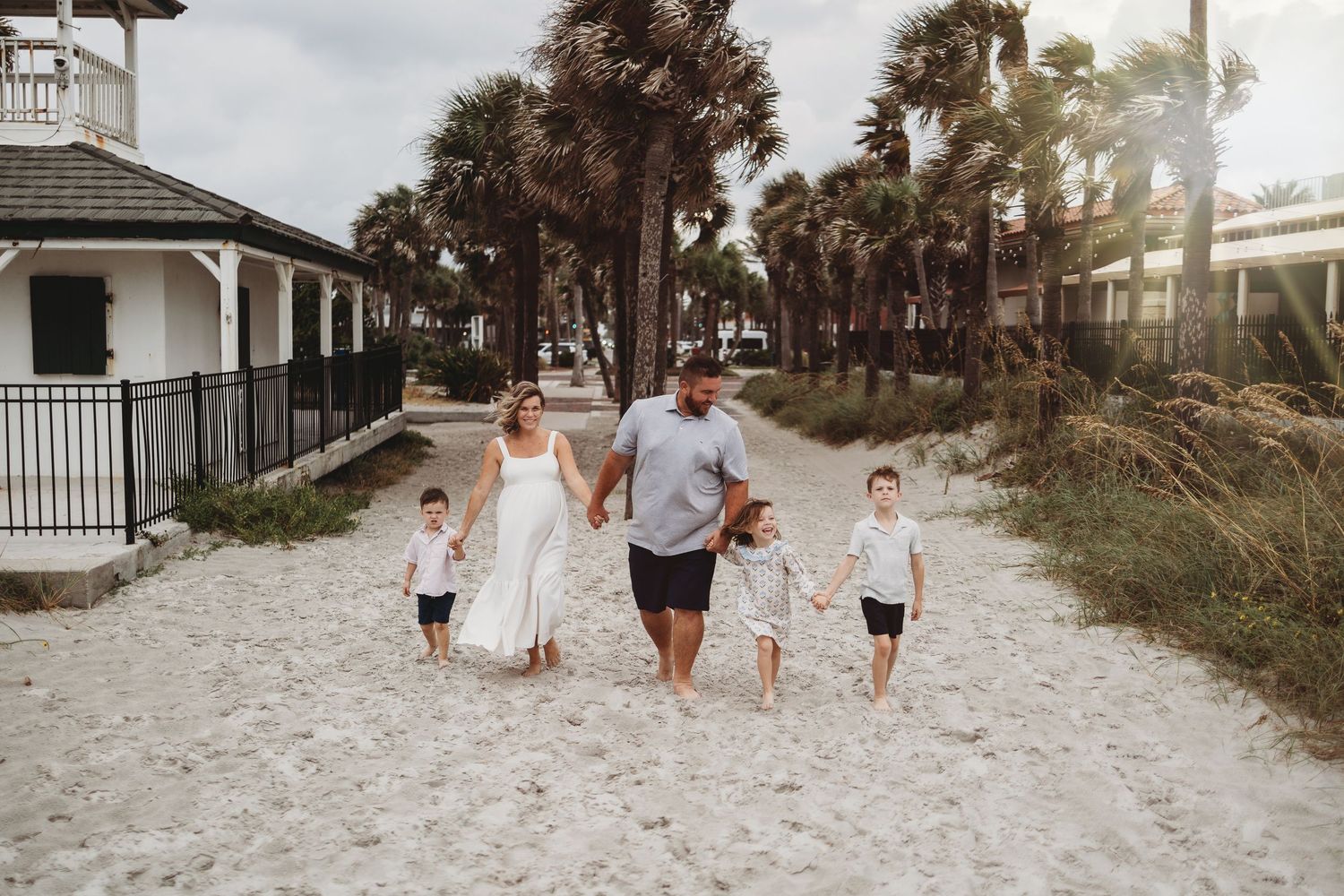 A family walks together along a white sandy beach path surrounded by palm trees and coastal vegetation.