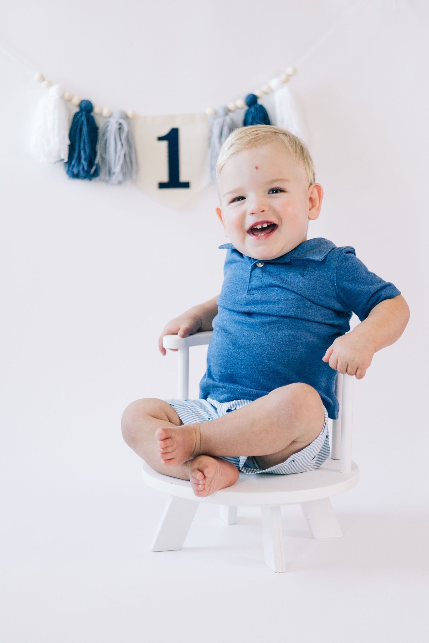 Baby in blue outfit sitting on white chair with number one decoration.
