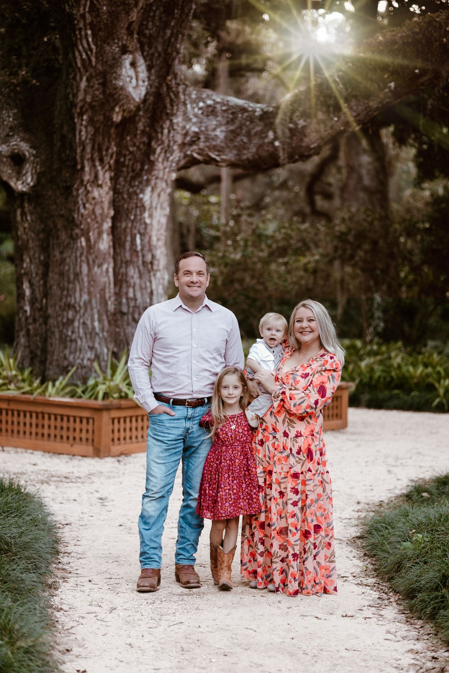 Family photo session on a sunlit white gravel path surrounded by lush trees.