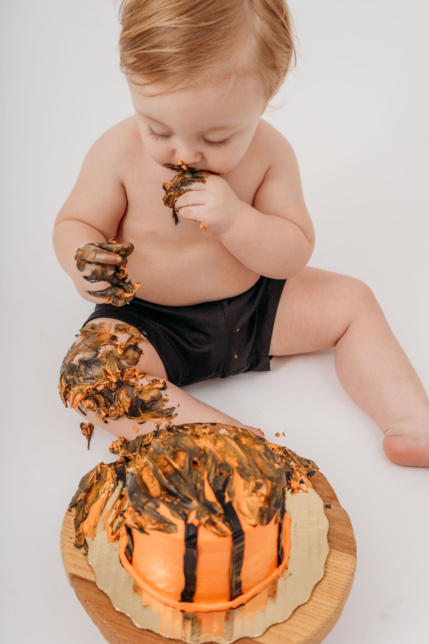 Baby in black diaper demolishing orange basketball cake during photoshoot.