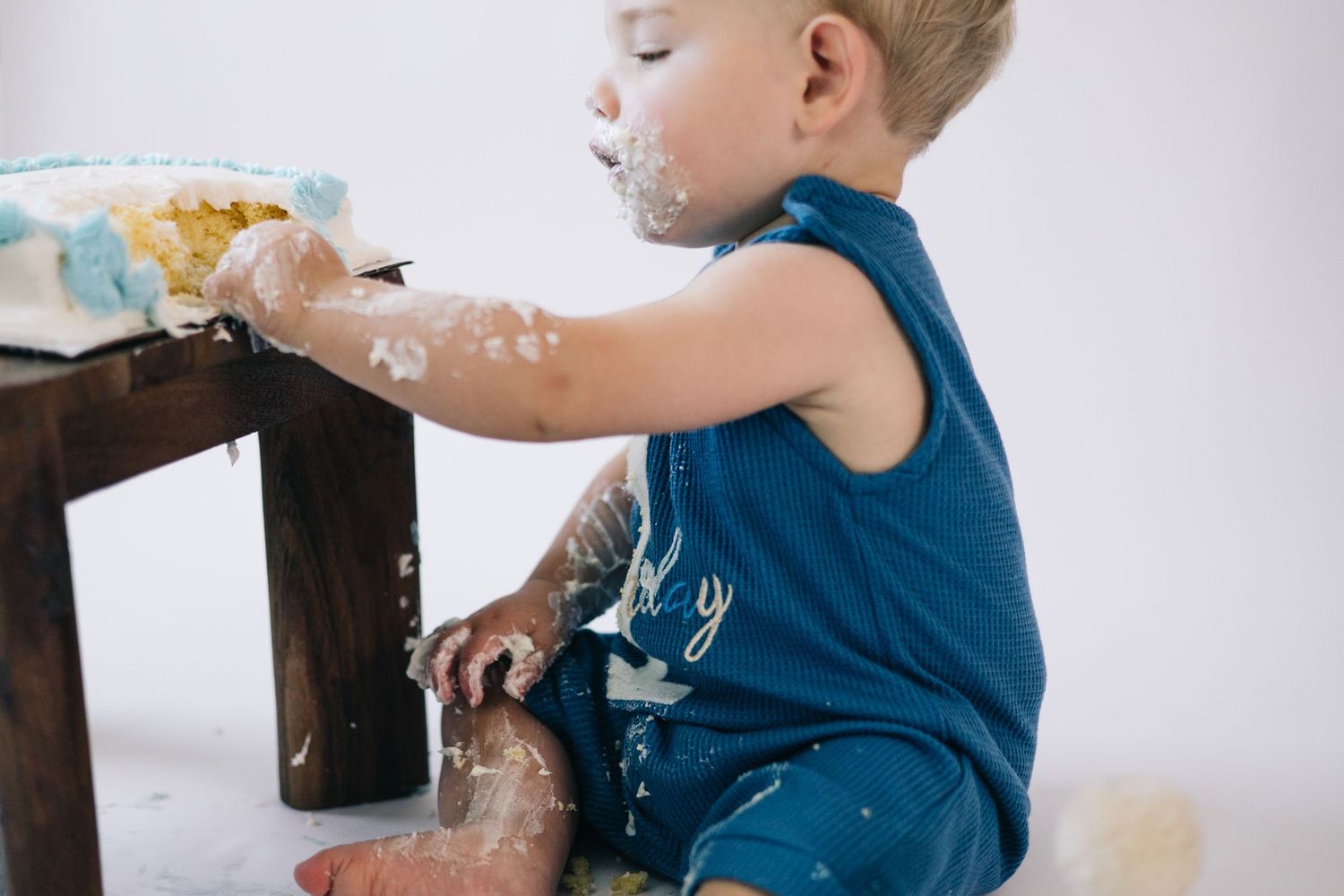 Messy cake smash photo session showing a baby in blue outfit playing with white frosting and chocolate cake.