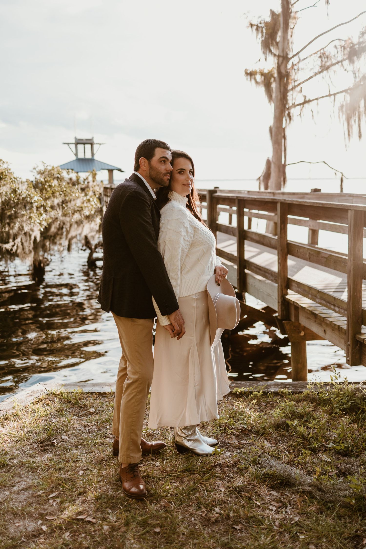 A romantic couple poses on a wooden dock overlooking a lake with Spanish moss hanging from trees at sunset.